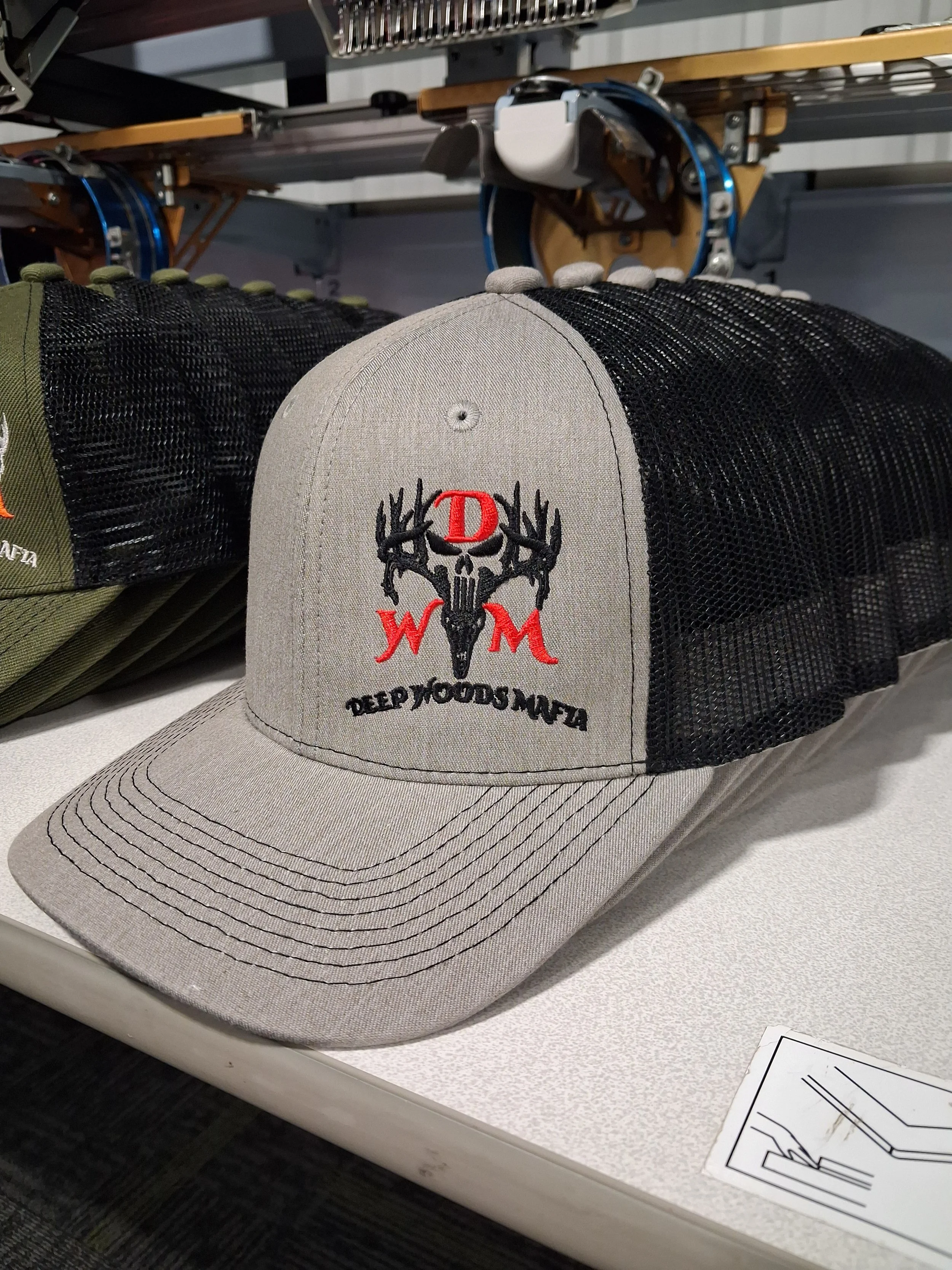 A beige baseball cap with a black deer skull logo and the words 'Deep Woods MTA' embroidered on the front, placed on a table next to other caps and a printed diagram.