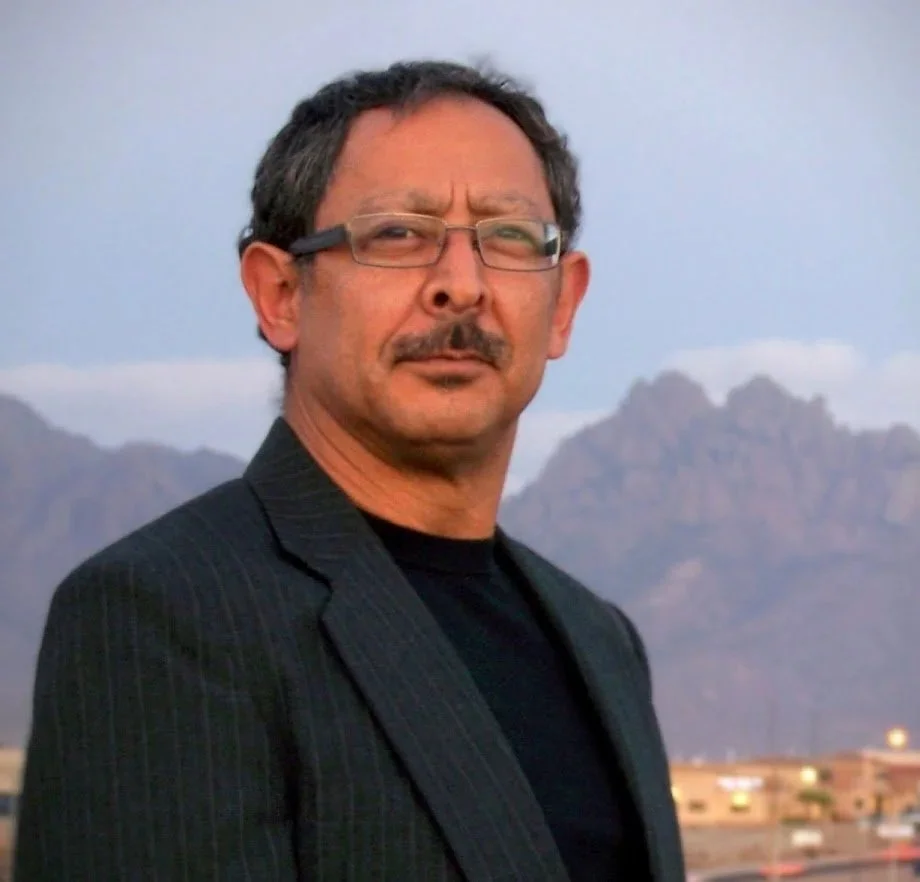 Man with glasses and mustache wearing a dark pinstripe suit and black shirt, standing outdoors with mountains in the background.