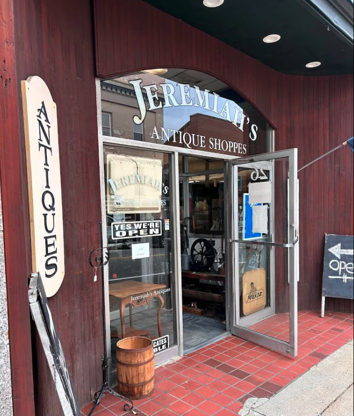 The entrance of Jeremiah's Antique Shoppes, a vintage store with glass double doors, a sign indicating it is open, and antique items visible inside. There is a vertical sign on the left with the word 'ANTIQUES' and a black chalkboard outside with an arrow pointing left and the word 'open' on it.