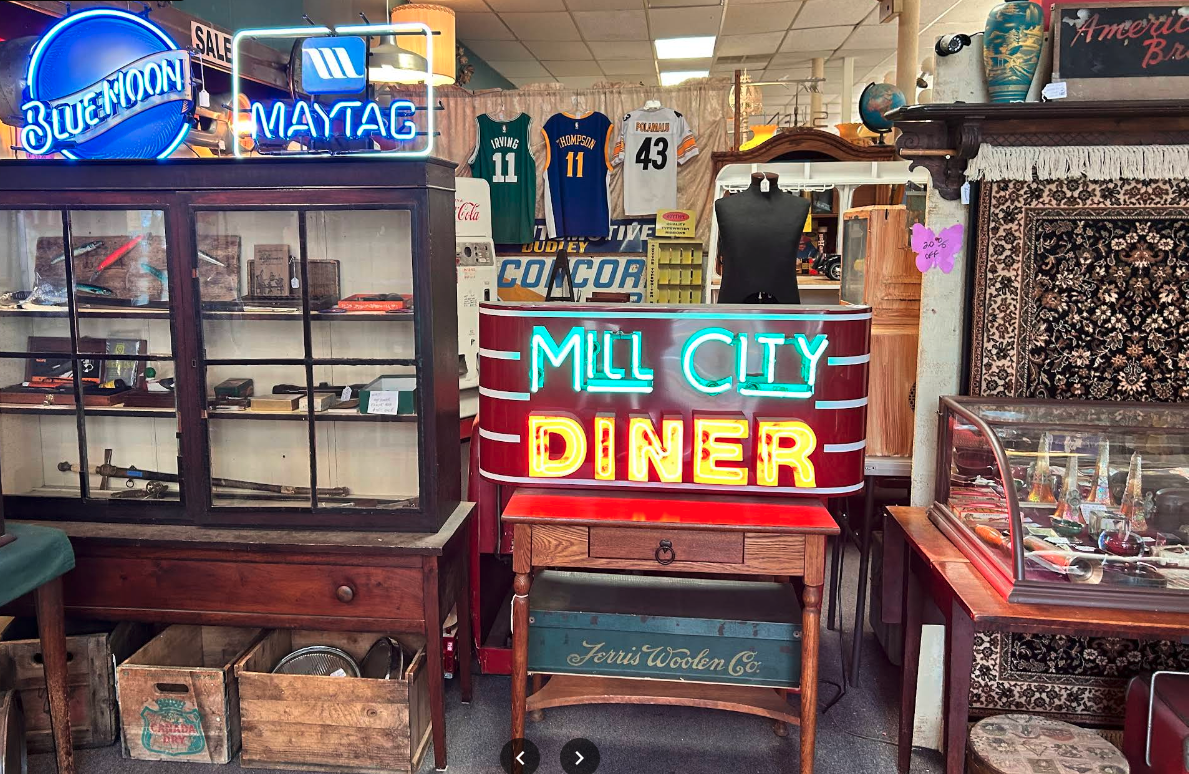 Interior of a vintage store with neon signs, sports jerseys, glass display cases with collectibles, and patterned rugs.
