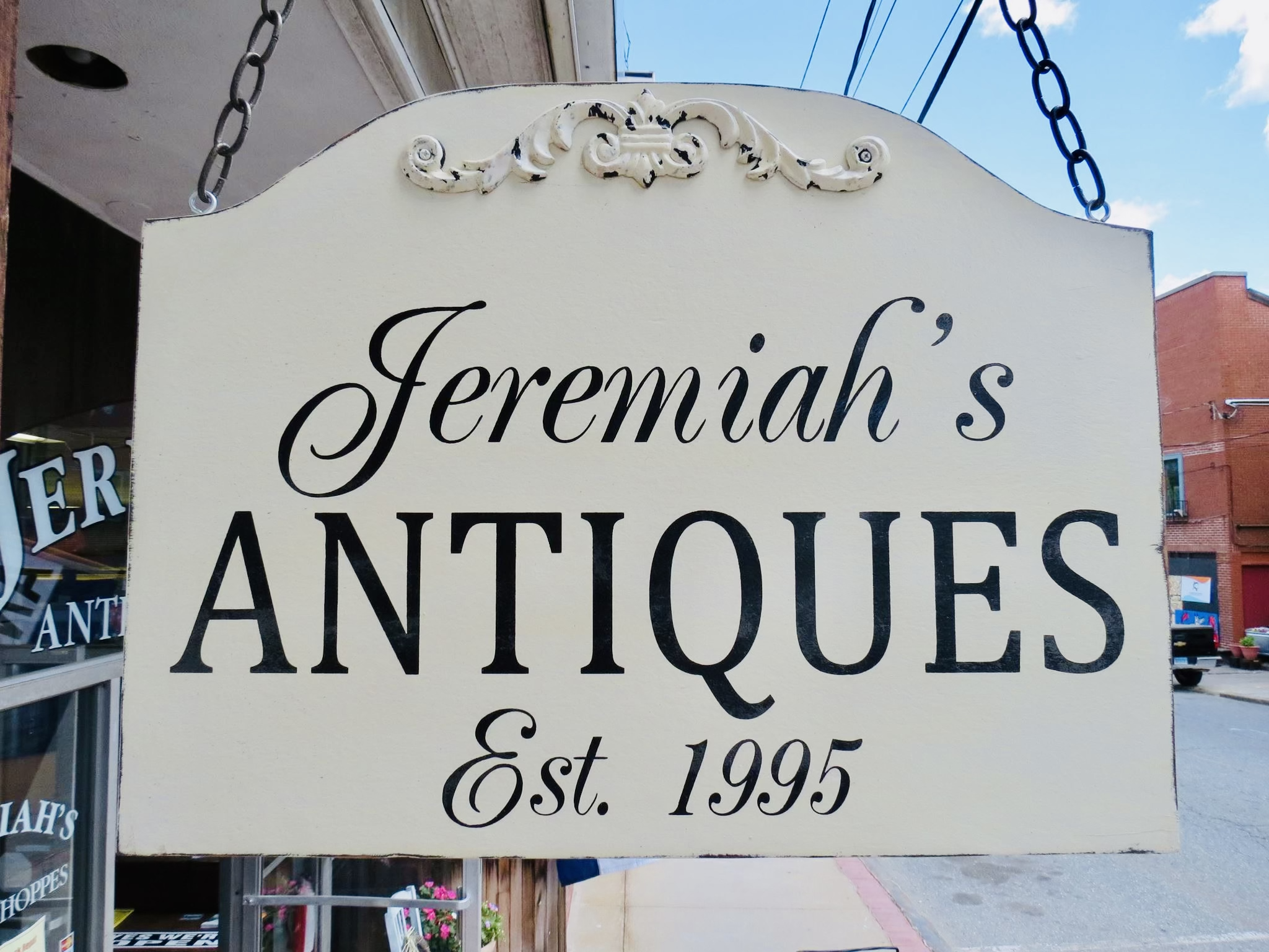 Sign for Jeremiah's Antiques established in 1995 hanging outside a shop in a small town, with a clear blue sky and brick buildings in the background.