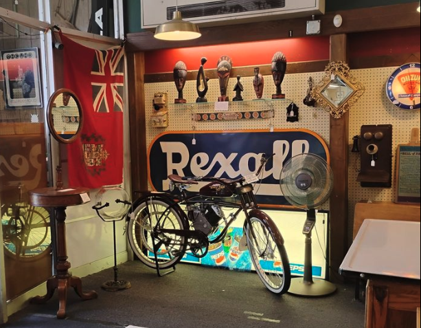 Vintage indoor display featuring a bicycle, Rexall sign, fan, flag, and various decorative items.