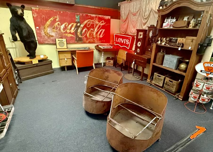 An antique shop with vintage items including a Coca-Cola sign, a bear statue, a Levi's neon sign, old furniture, containers, and rusted metal tub-like carts.