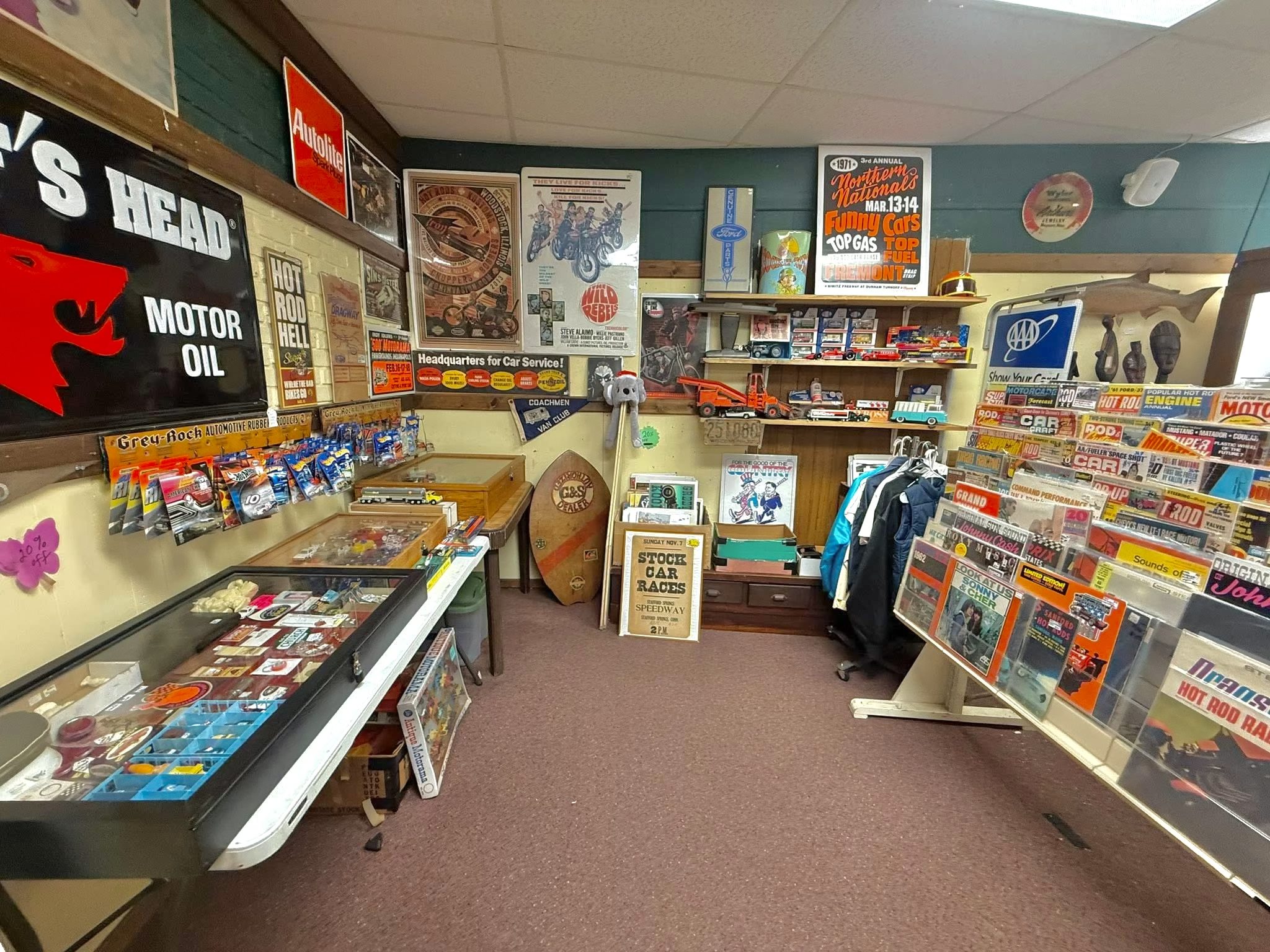 The image shows a vintage automotive collectibles store with shelves of magazines and posters, a display case with small car models, and various automotive memorabilia on the walls.