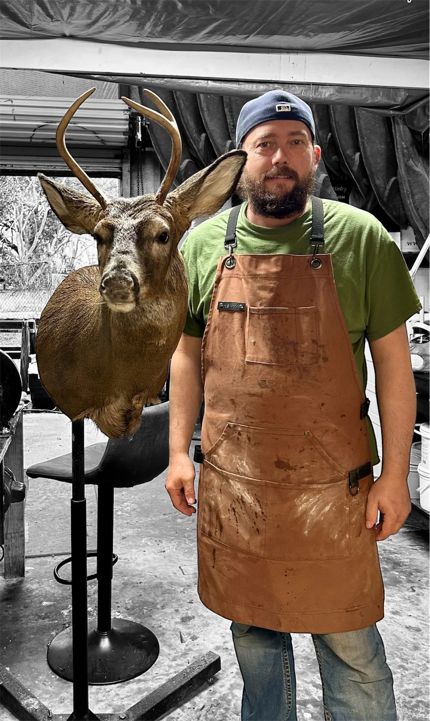 A man with a beard wearing a green t-shirt, baseball cap, and a stained leather apron, standing next to a mounted taxidermy deer head in a workshop or garage setting.