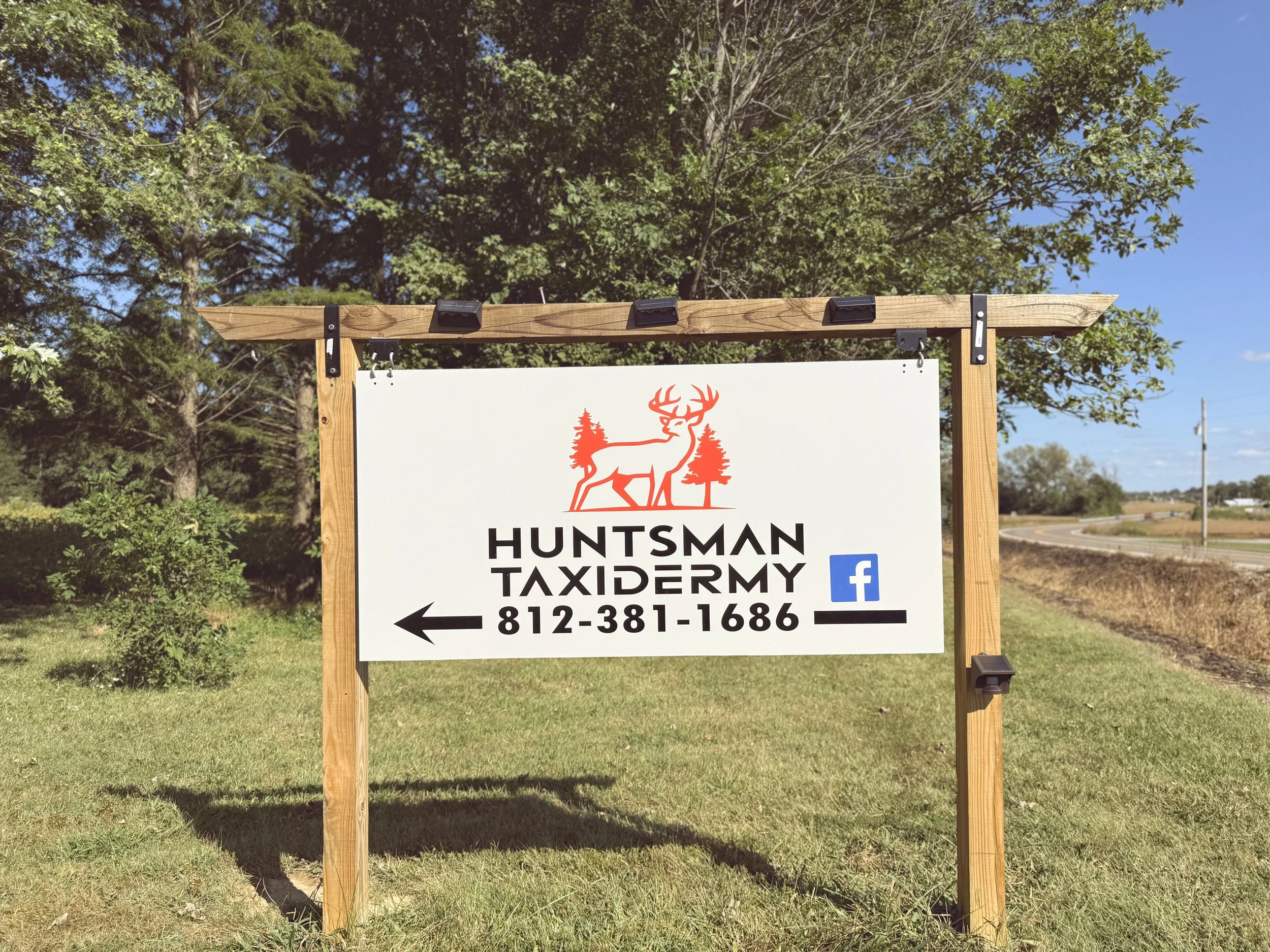 Sign for Huntsman Taxidermy with a red deer silhouette and trees logo, phone number 812-381-1686, pointing left, with Facebook icon, outdoor setting with grass, trees, and a blue sky.