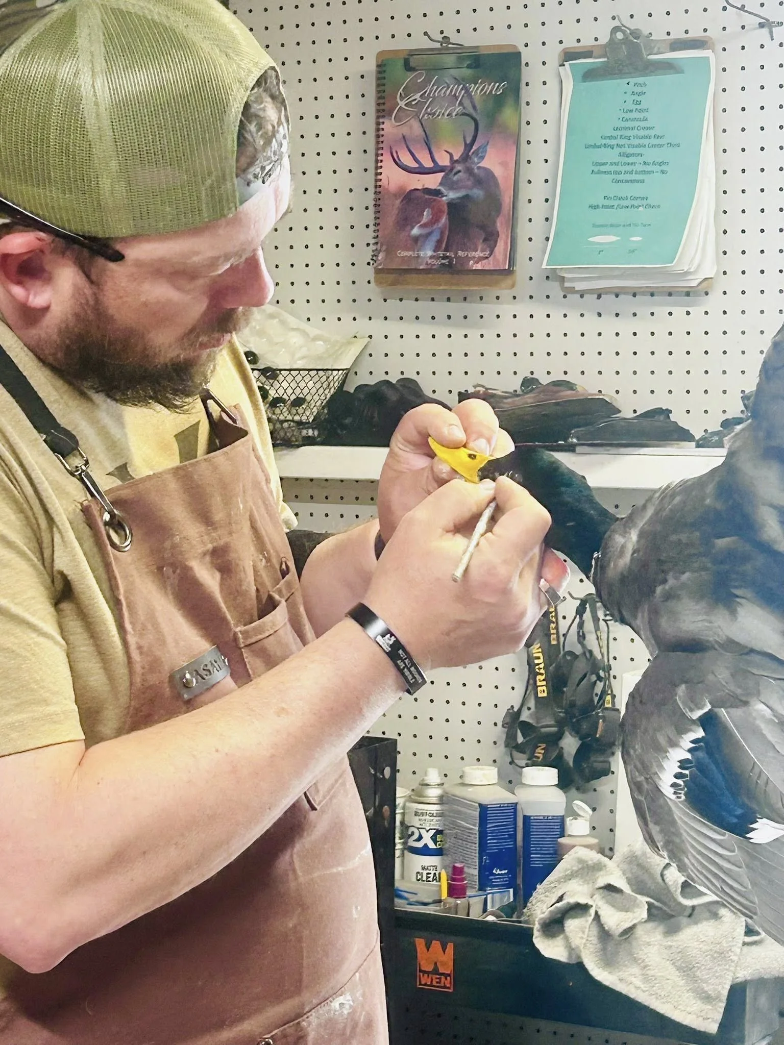 A man wearing a green cap, glasses, a tan t-shirt, and a brown apron, painting the beak of a mounted bird in a workshop with various tools and supplies, including a small poster with a deer on the wall.