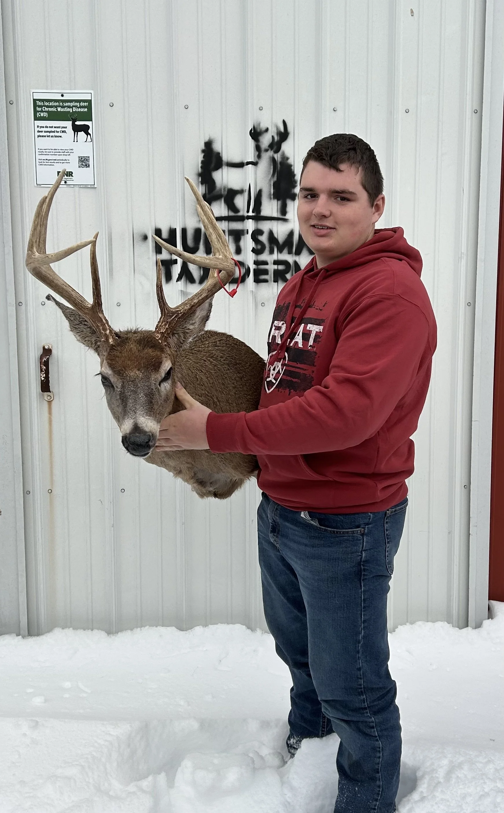 A young man in a red hoodie and jeans standing on snow while holding a mounted deer head with antlers