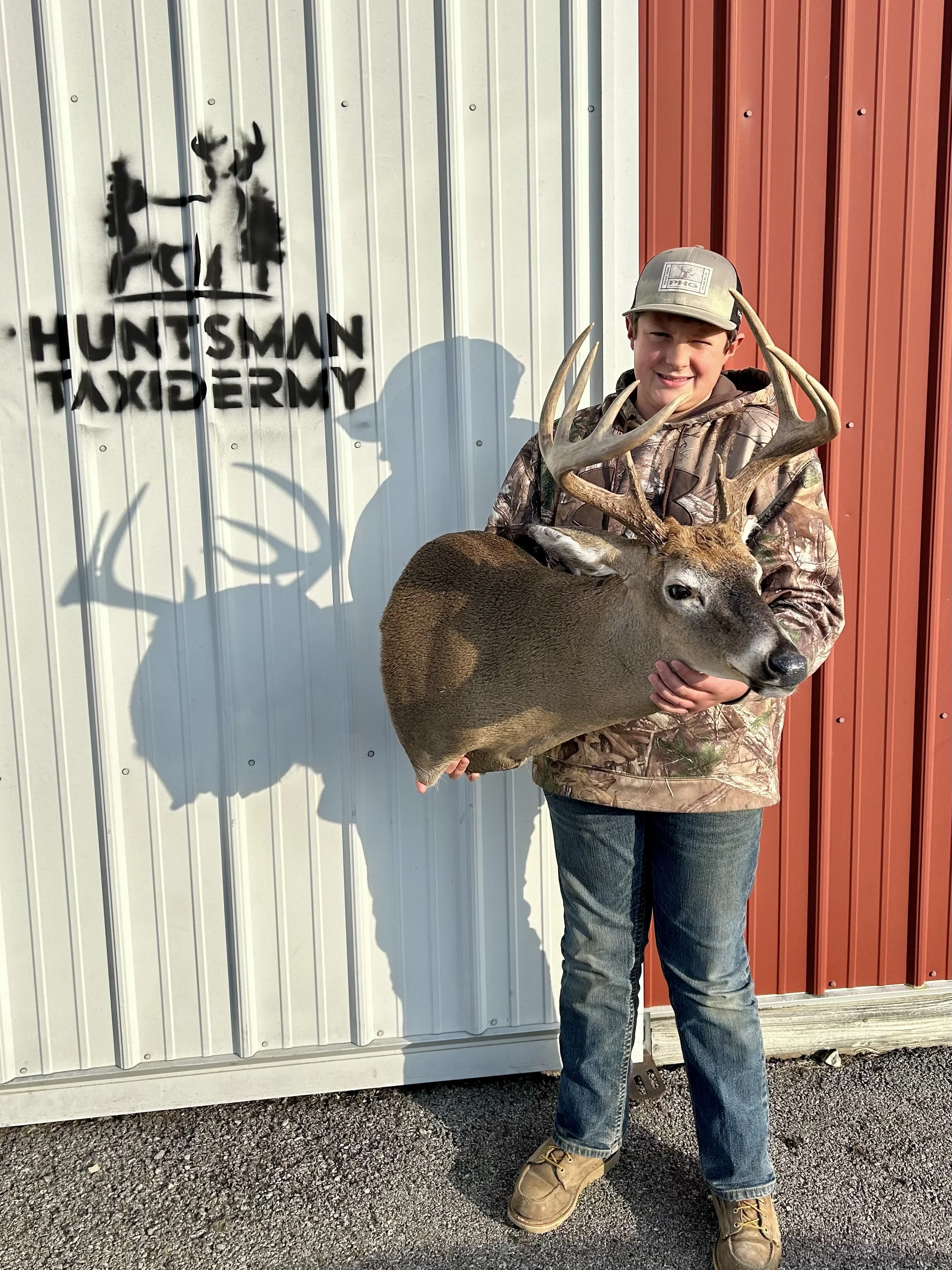 A young boy in camouflage jacket and jeans holds a large deer with antlers, standing outside a building with white and red metal siding and a 'Huntsman Taxidermy' sign.