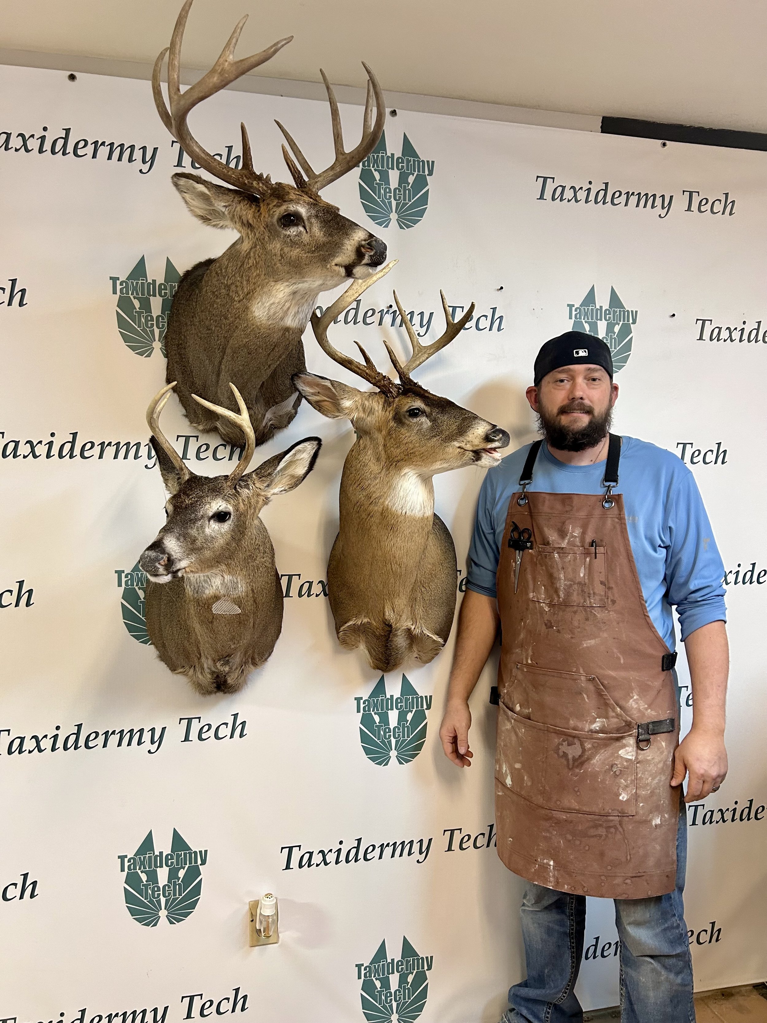 A man in a blue shirt and brown apron standing next to three mounted deer heads on a wall. The background has the logo and name 'Taxidermy Tech' repeated multiple times.