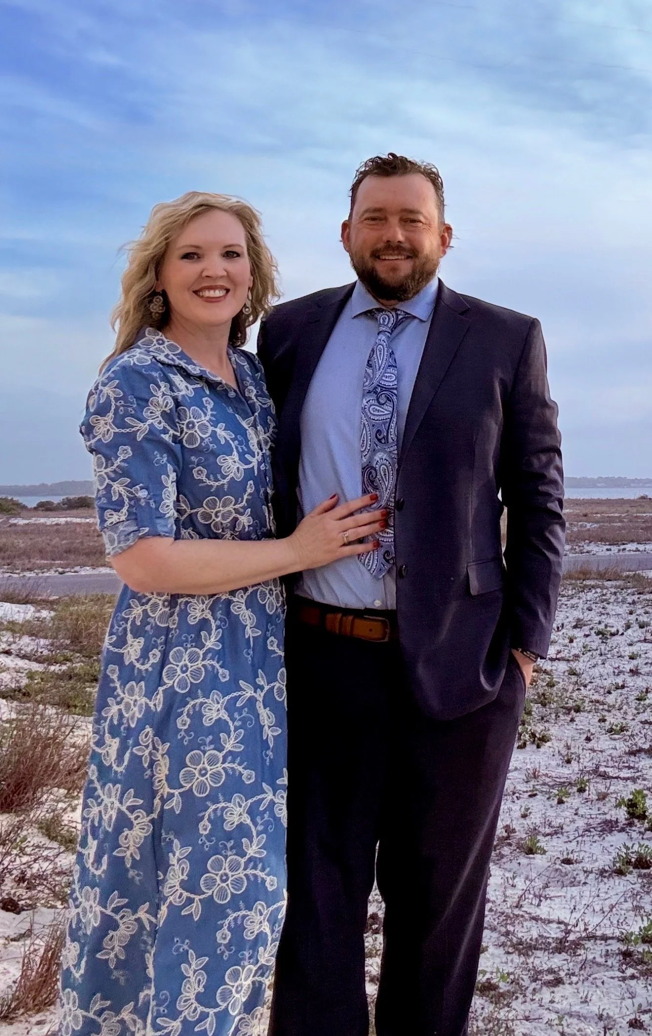 A smiling woman and man standing outdoors on a beach with dunes and water in the background, dressed in semi-formal attire.