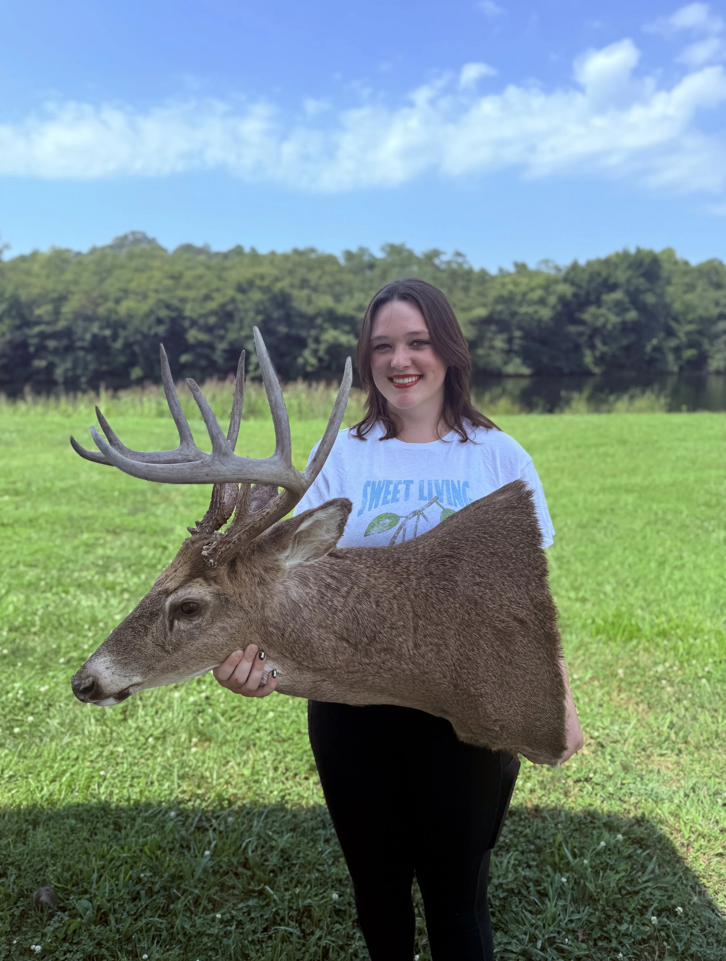 Young woman with dark hair, smiling, holding a large deer head with antlers outdoors on a grassy field near a lake, with trees and partly cloudy sky in the background.