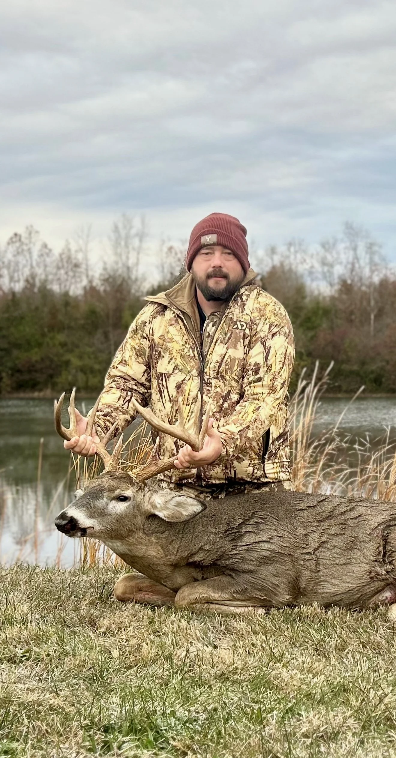 A man in a camouflage jacket and maroon beanie holding a large deer by its antlers near a riverbank with trees and grass in the background.