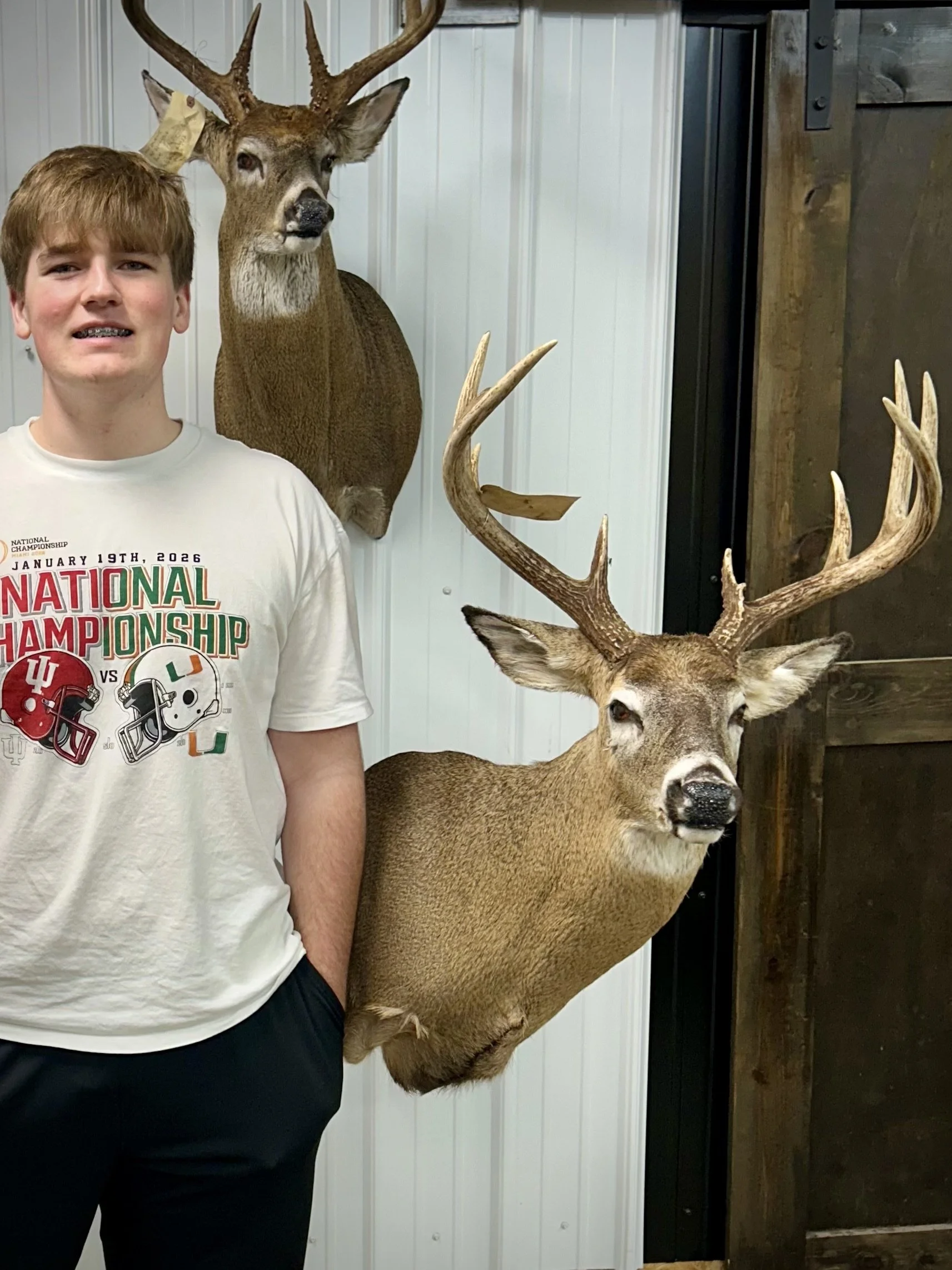 A young boy standing next to two mounted deer heads on a wall in an indoor setting.