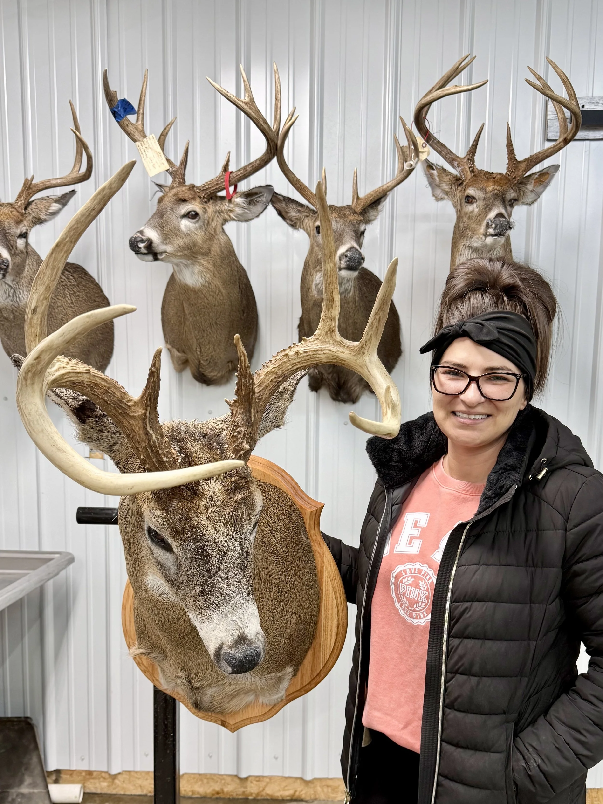A woman with glasses and a black headband poses next to a mounted deer head with large antlers, surrounded by five other mounted deer heads on a vertical metal-paneled wall.