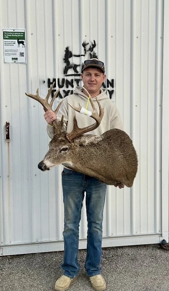 Man holding a large deer with antlers, standing in front of a white metal building with hunting sign.