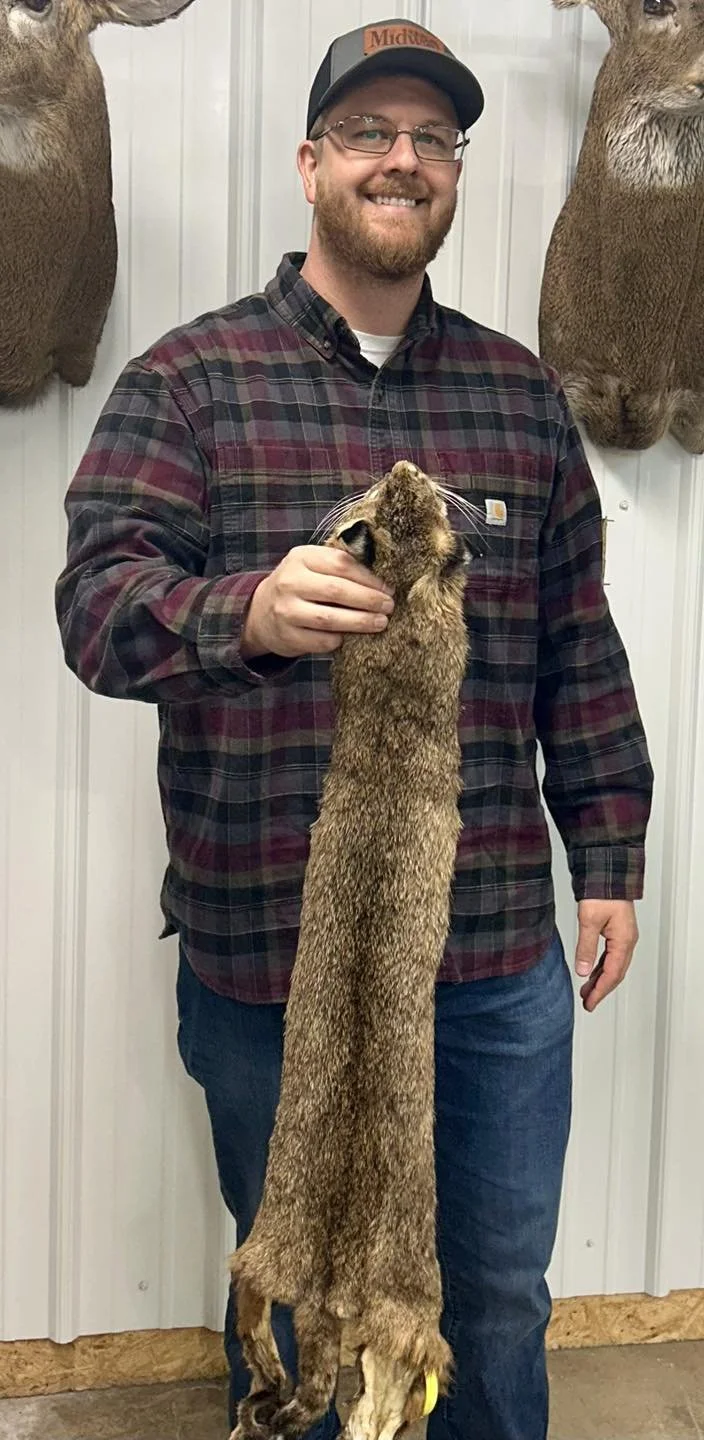 A man with glasses, wearing a plaid shirt and baseball cap, smiling and holding a large, long-bodied rodent or similar animal in front of a wall with mounted deer heads.