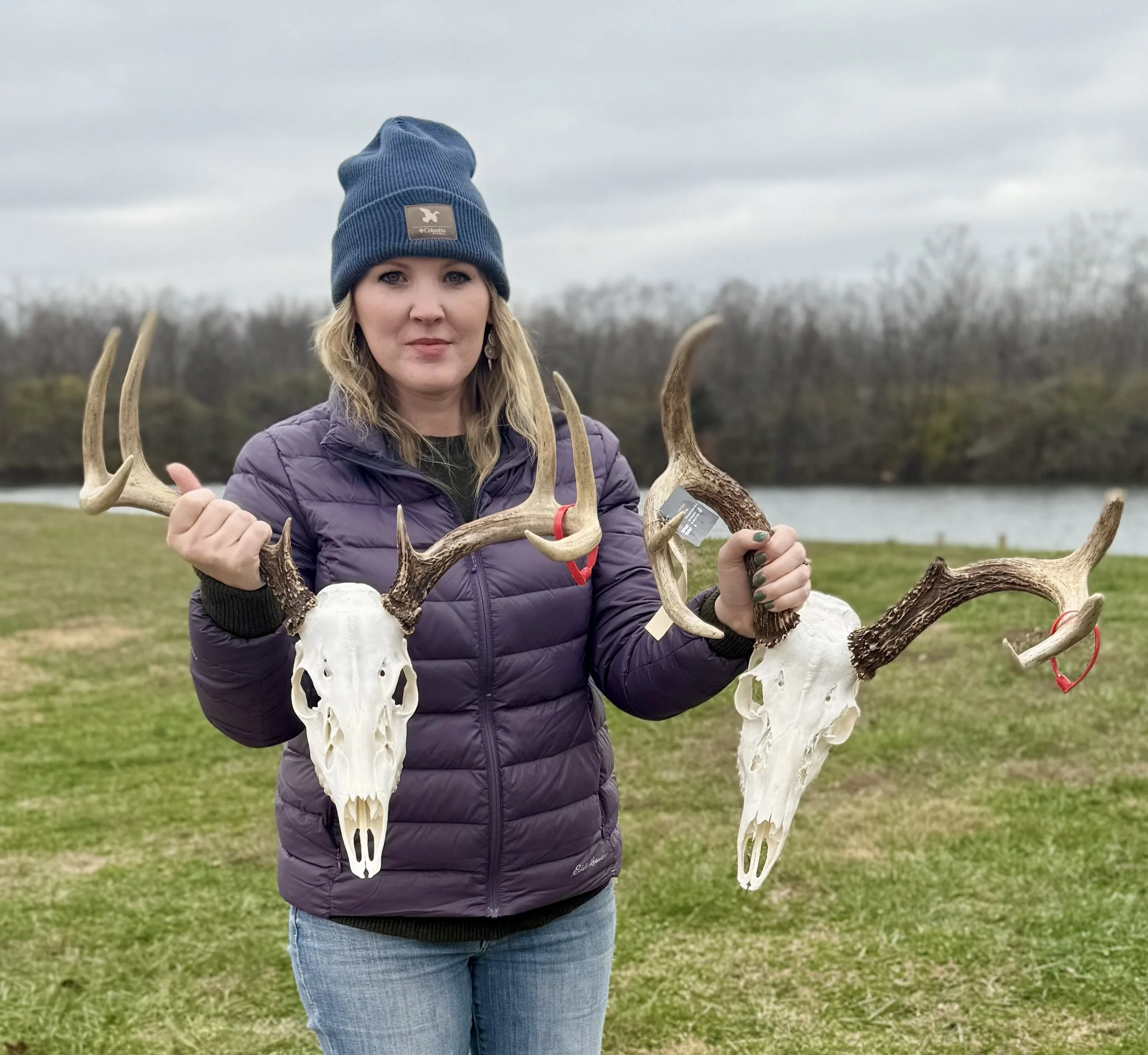 Woman in purple jacket and blue beanie holding two deer skulls with antlers outdoors on a grassy area near a body of water.