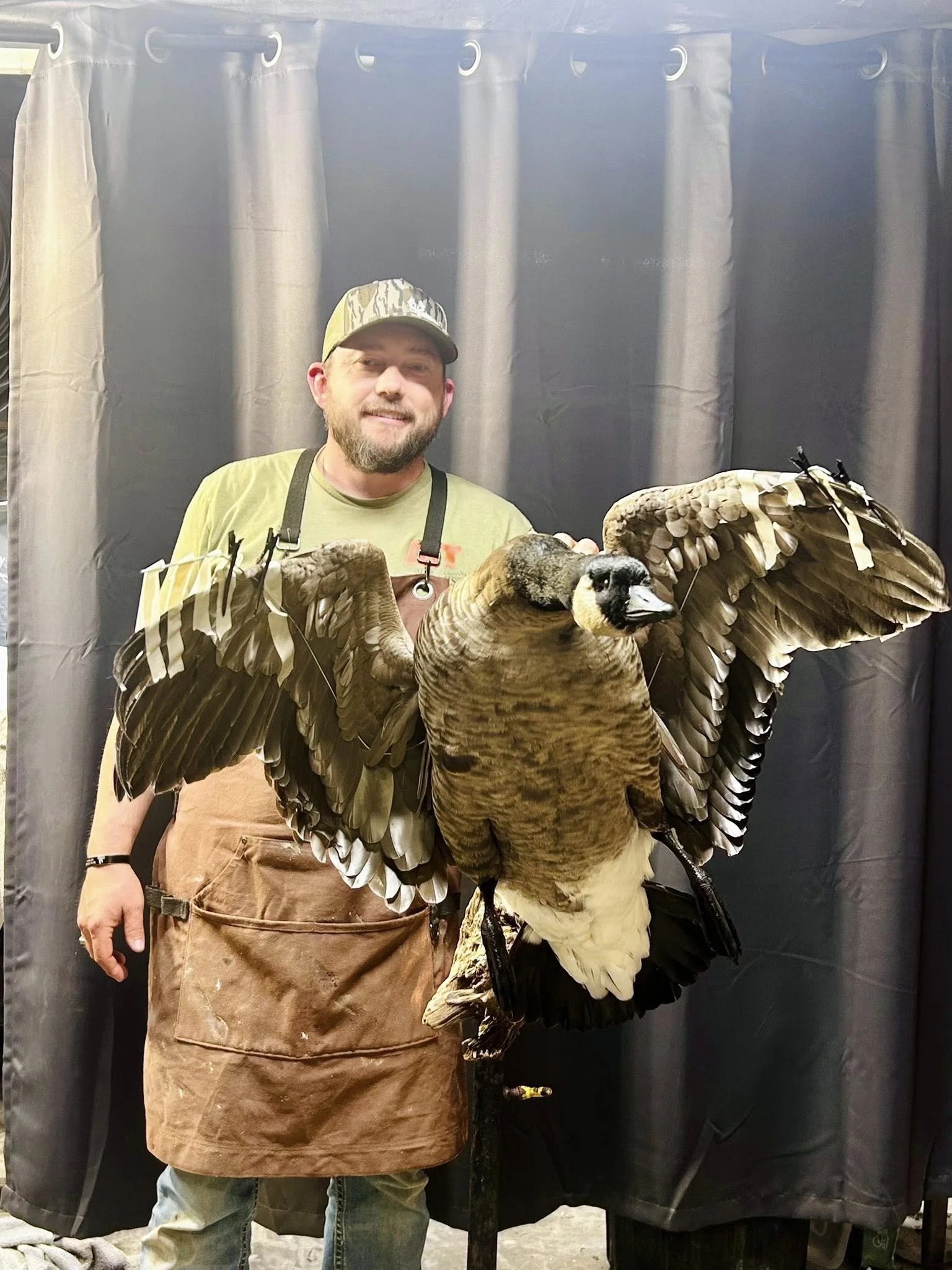 A man with a beard wearing a camouflage cap, a green shirt, and a brown apron stands smiling next to a mounted Canadian goose with its wings spread.