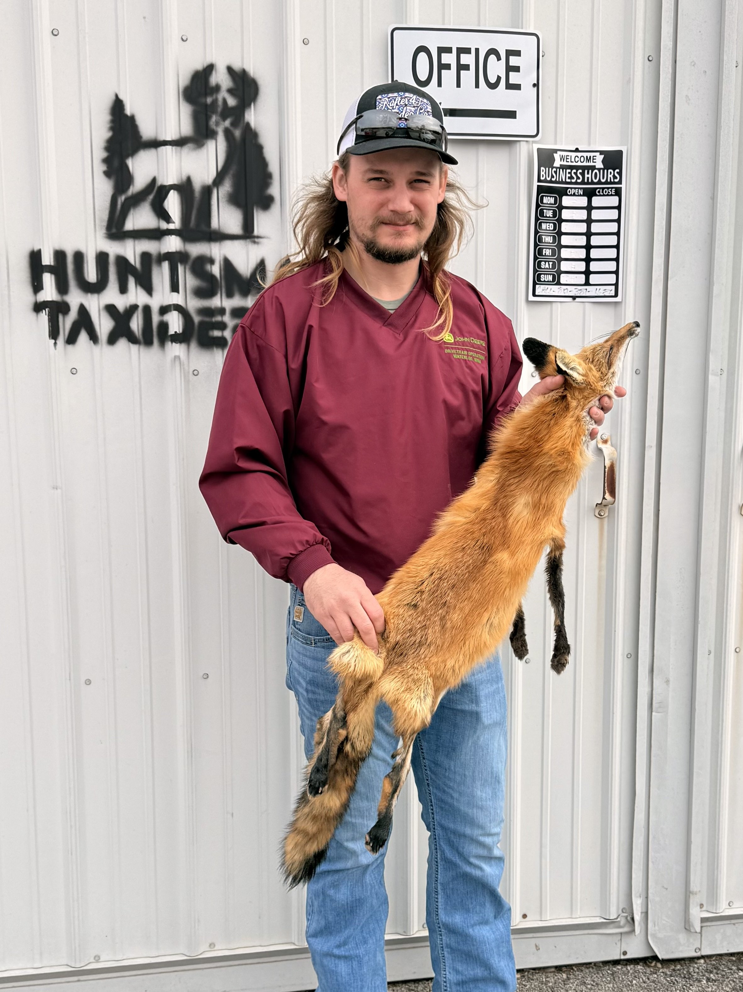 A man with long hair, wearing a maroon jacket and a cap, holding a small fox named Hunt. The background features a wall with the logo and name of Hunt's Taxi, an office sign, and a business hours schedule.