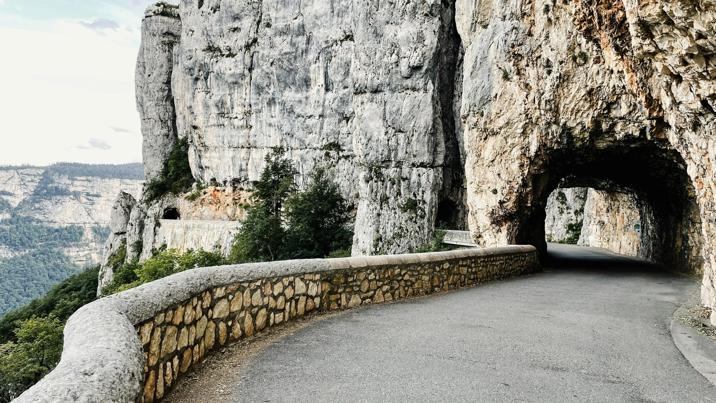 Une route serpentant à travers un tunnel naturel formé par des rochers dans une zone montagneuse, avec des falaises rocheuses à gauche et une barrière en pierre à gauche de la route.