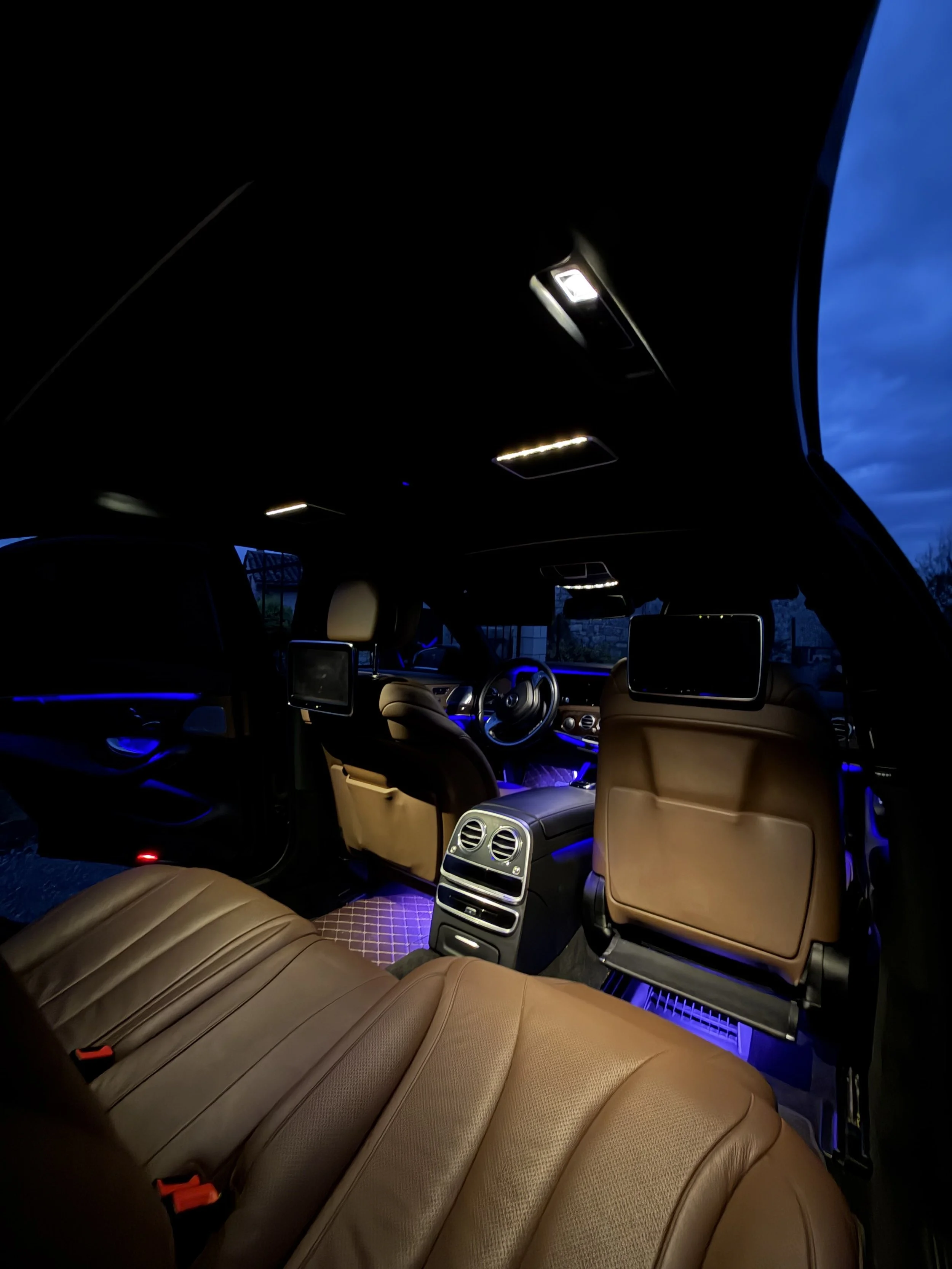 Intérieur luxueux d'une voiture avec sièges en cuir beige, éclairage d'ambiance violet, écrans de divertissement et tableau de bord moderne.