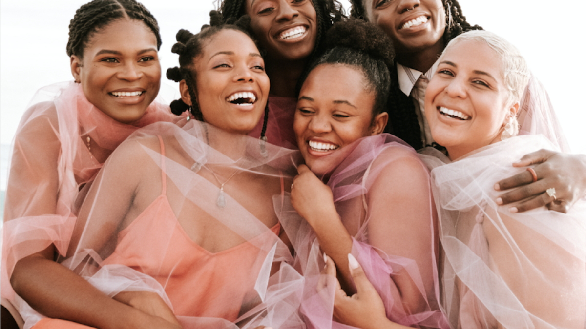 Group of seven women smiling and hugging each other, wearing soft pink, peach, and white tulle dresses.
