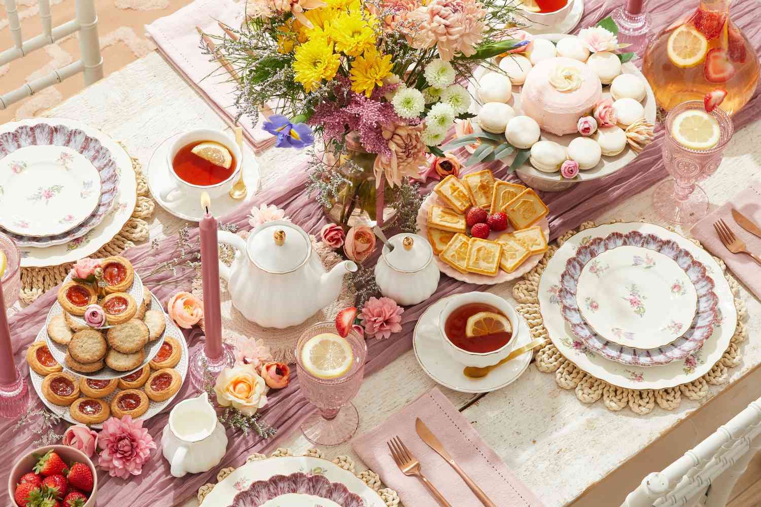 A tablescape with a pink tablecloth, floral centerpiece, teacups with tea, an array of desserts including cookies, scones, macarons, strawberries, and lemon slices, vintage floral plates, and pink candles for a tea party.