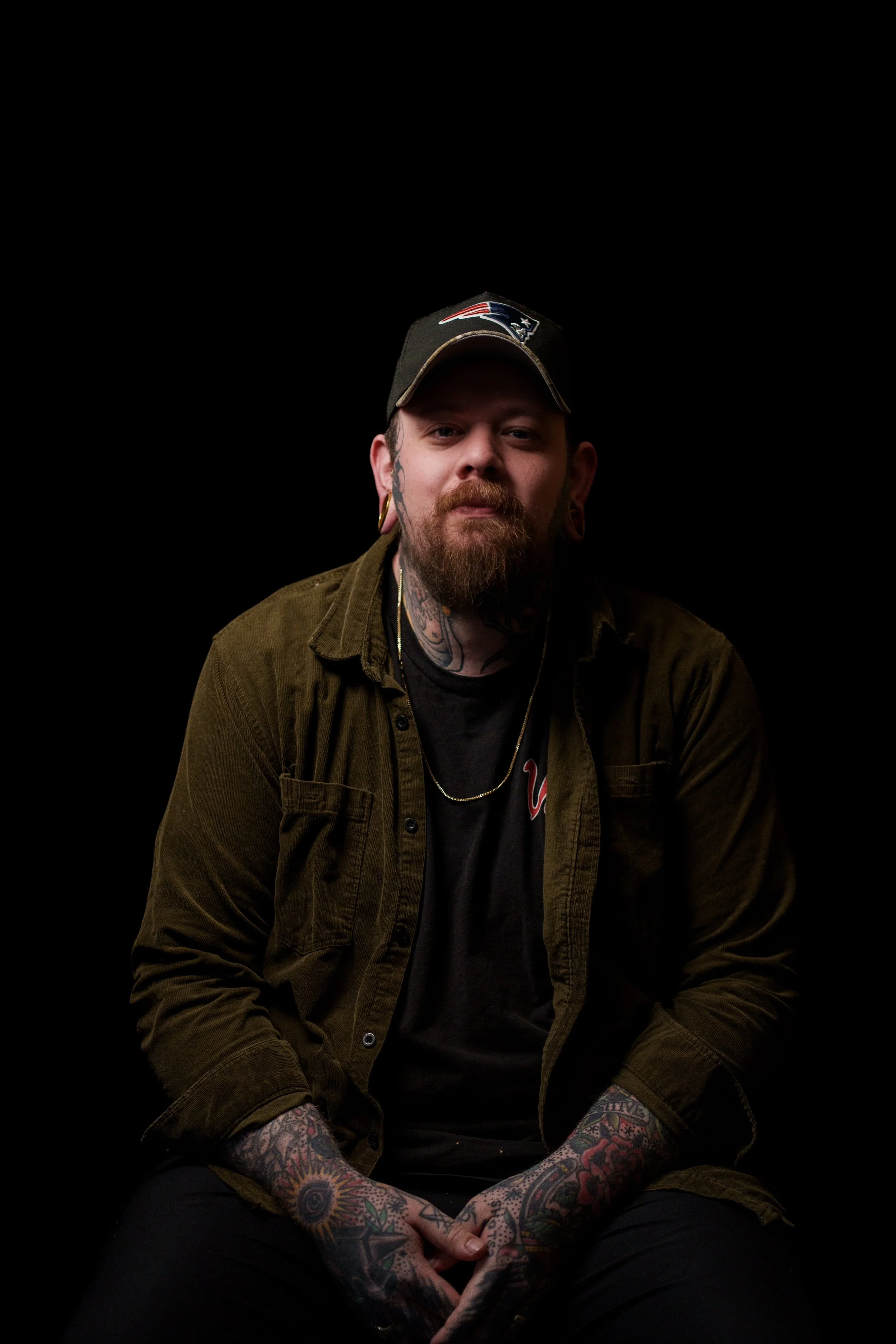 A man with tattoos on his arms and neck wearing a green jacket and a baseball cap sitting against a black background.