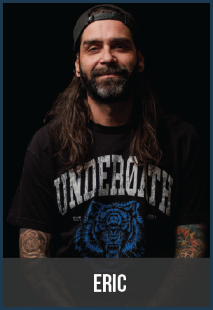 Portrait of a man with long hair, beard, wearing a black cap backward and a black T-shirt with blue and white text and graphic, standing against a dark background.