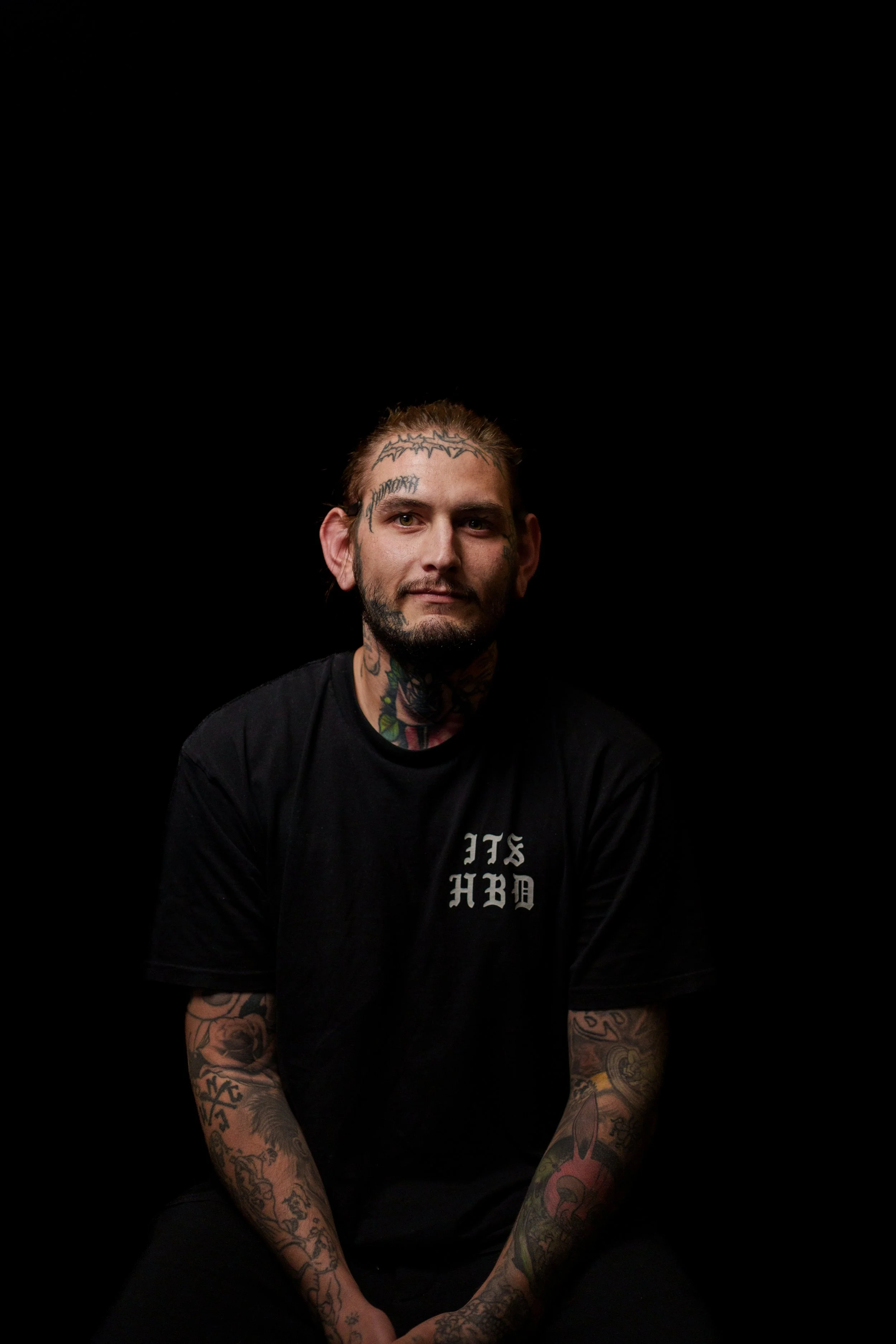 A man with tattoos on his face, neck, and arms sitting against a black background, wearing a black shirt.