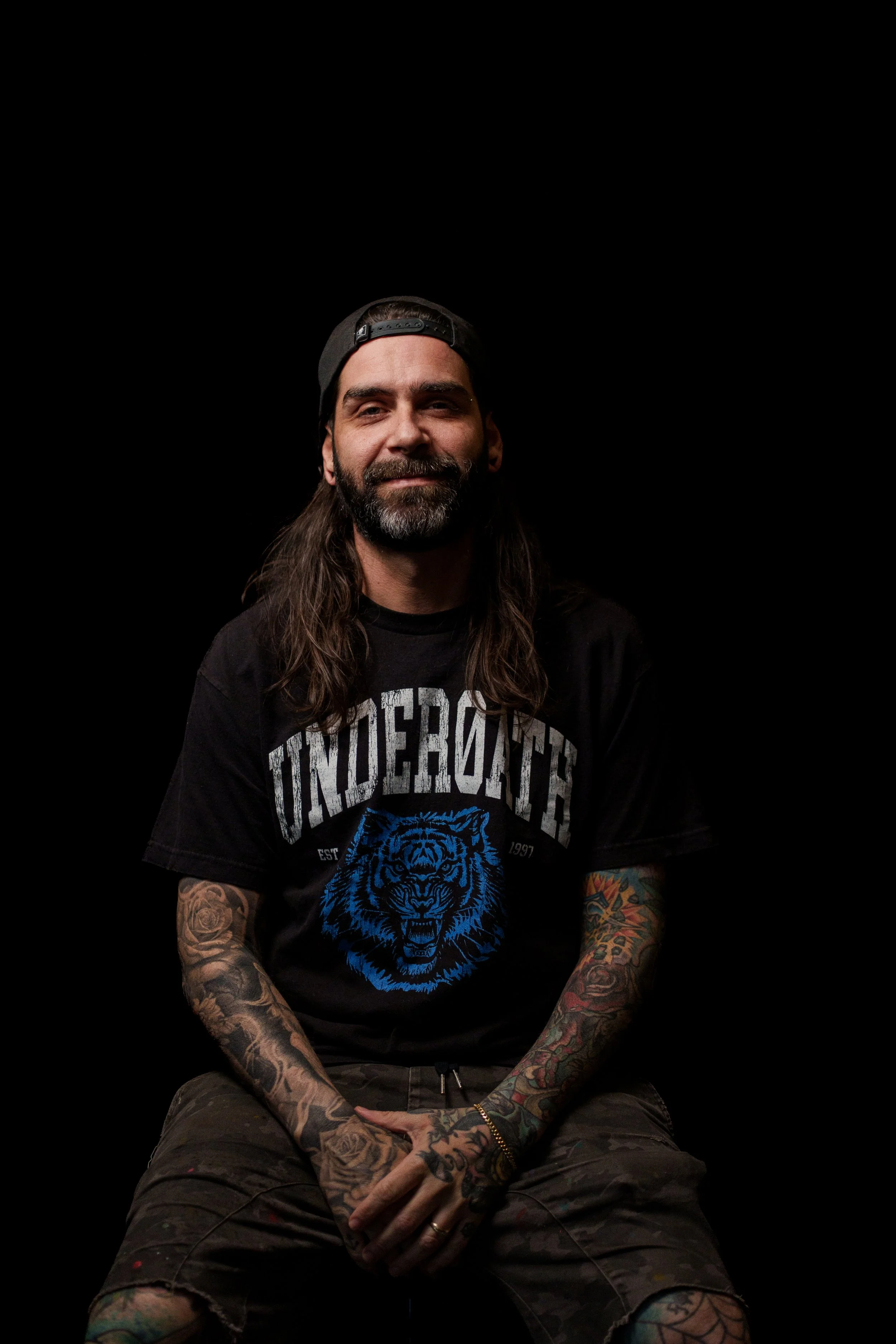 A man with long hair and tattoos on his arms, wearing a black cap backwards and a black T-shirt with a blue roaring tiger graphic and the word 'UNDERCATOR' on it, sitting against a black background.