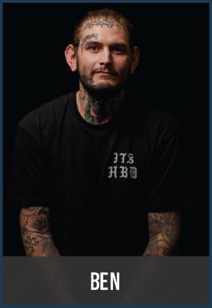 Portrait of a man with tattoos, dark hair, and a beard, wearing a black T-shirt with white text, against a black background