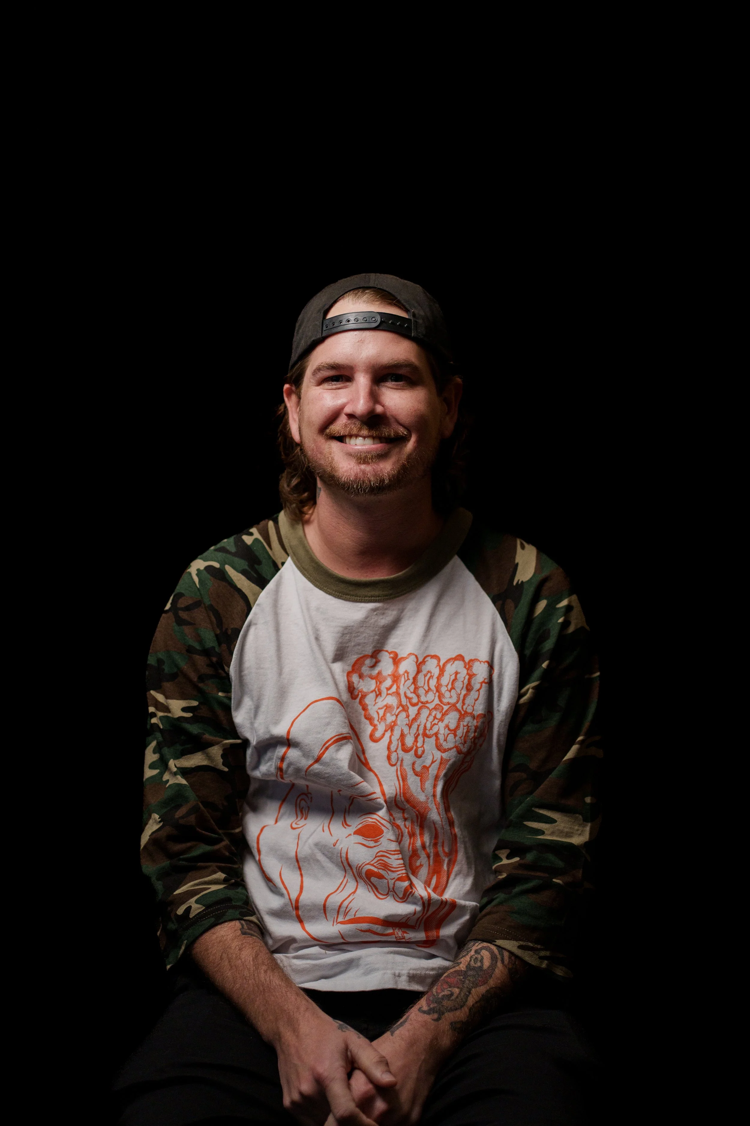 A smiling man with long hair, beard, and mustache, wearing a black backwards cap, a white T-shirt with orange graphic art, camouflage patterned sleeves, tattoos on his left arm and right wrist, sitting against a plain black background.