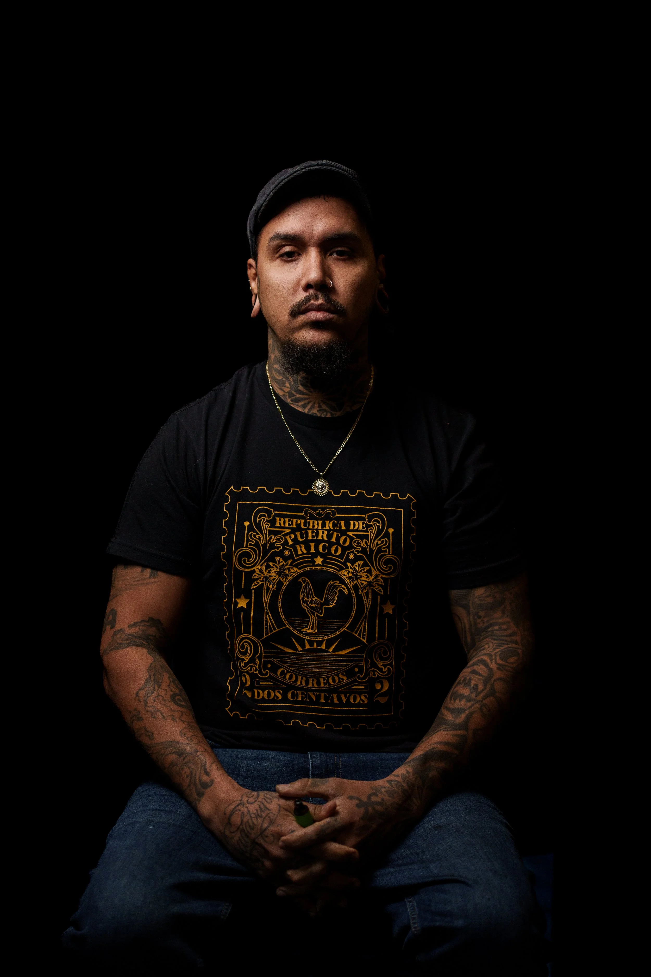 Portrait of a man with tattoos, piercings, and a beard, wearing a black cap and a black T-shirt with a gold design, seated against a black background.