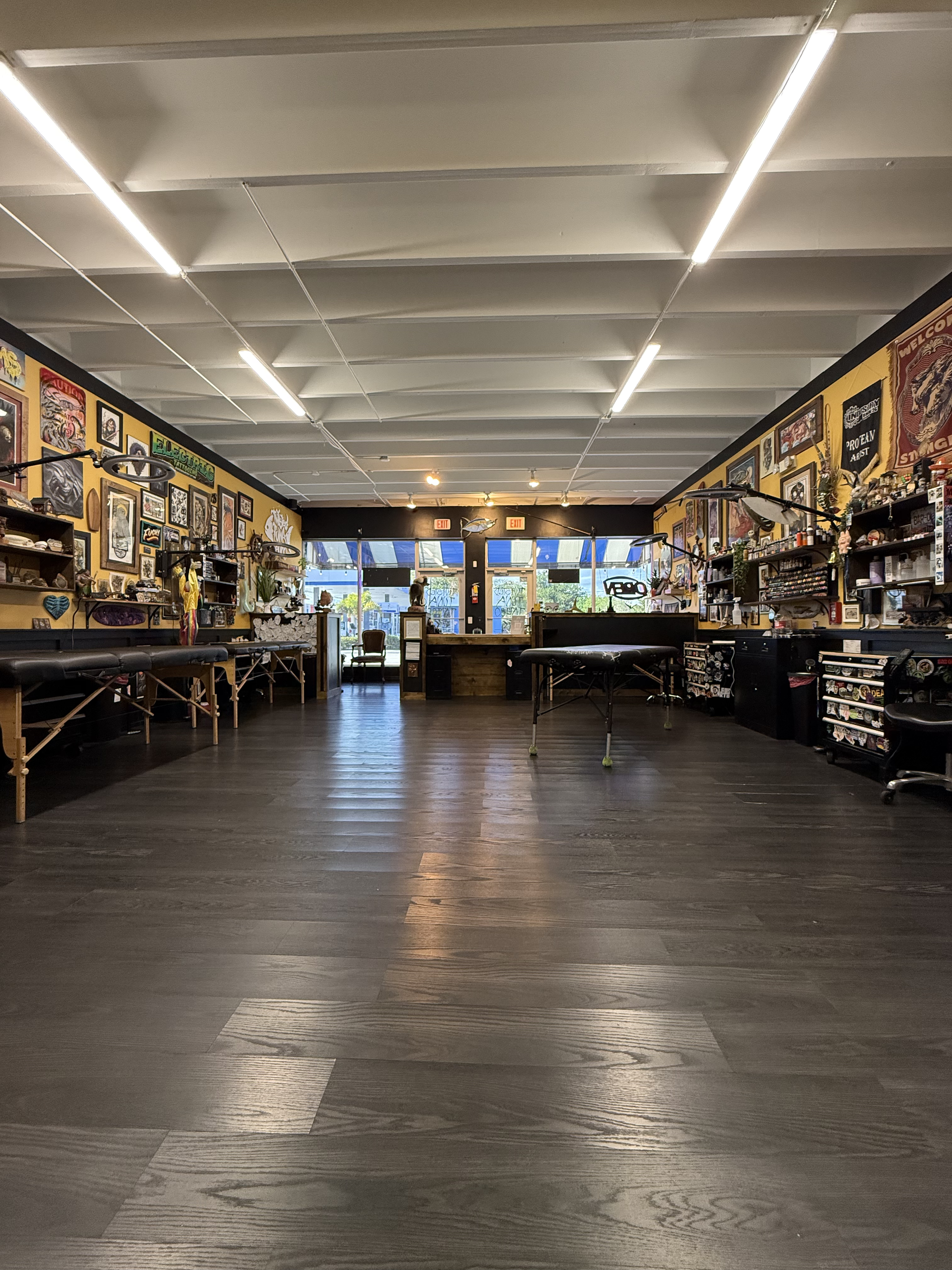 Clean tattoo shop with dark wooden floors, yellow and black walls decorated with art and posters, and shelves with art supplies. There are adjustable lamps over work tables, and a front window with a door, showing parked cars outside.