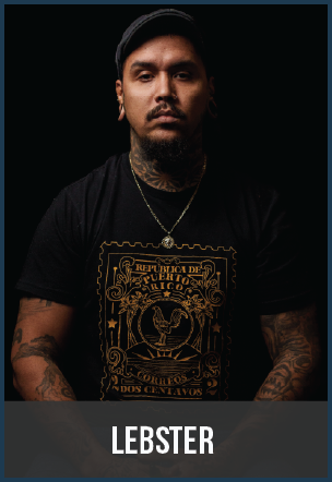 Portrait of a man with tattoos wearing a black T-shirt and a necklace, against a dark background.