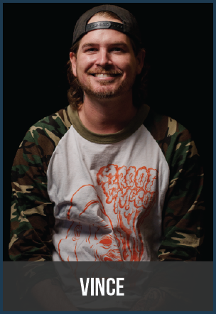 Smiling man wearing a baseball cap backwards, camo sleeves shirt, and a graphic t-shirt, against a dark background, with a caption 'Vince'.