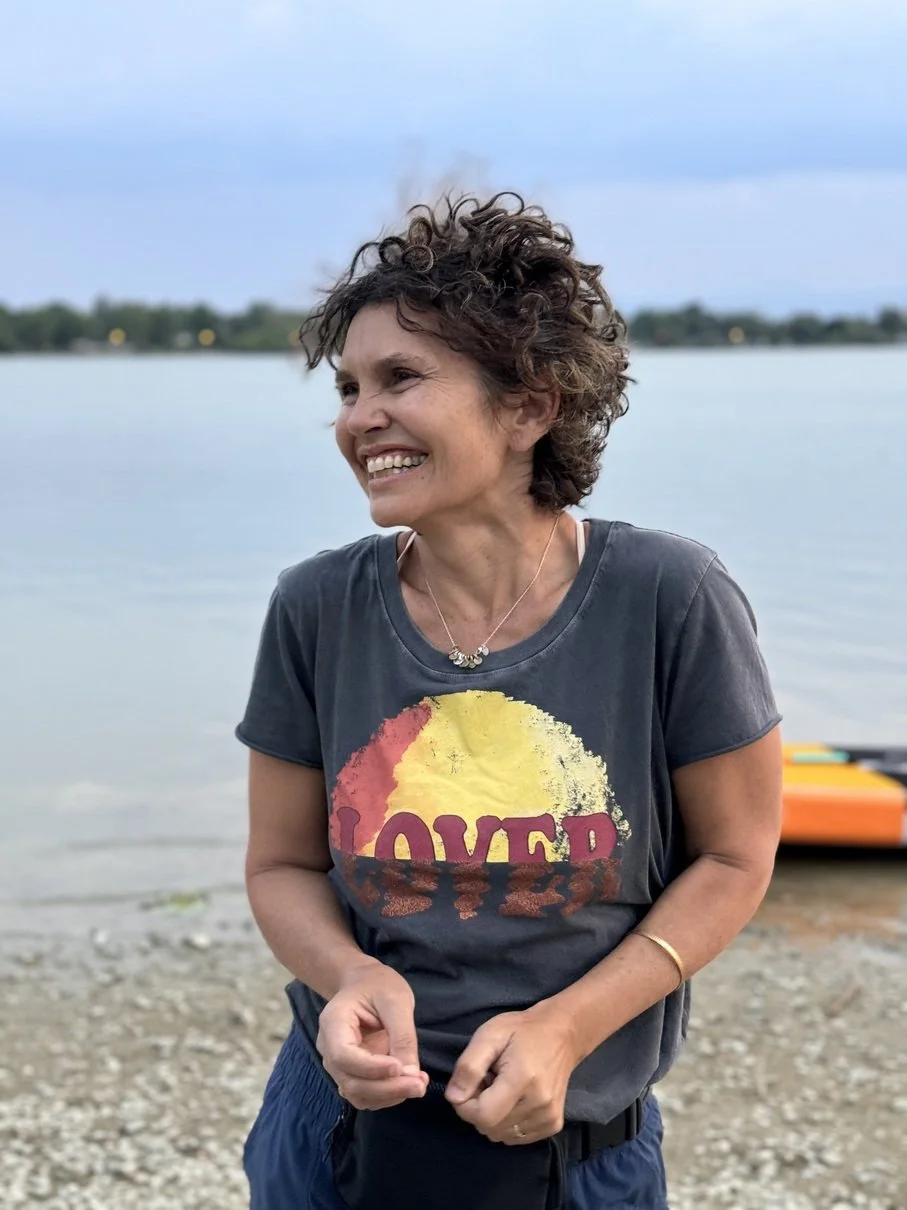 A woman with curly hair smiling while standing by a lake, wearing a gray T-shirt with a colorful sunset design and the word "LOVED" on it, with a paddleboard visible in the background.