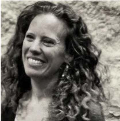 A woman with curly hair smiling outdoors against a stone wall.