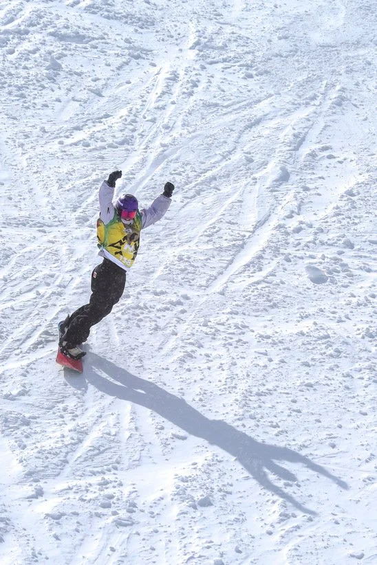 A person snowboarding down a snowy slope during daytime, wearing colorful winter gear and gloves, with their arms raised in excitement.