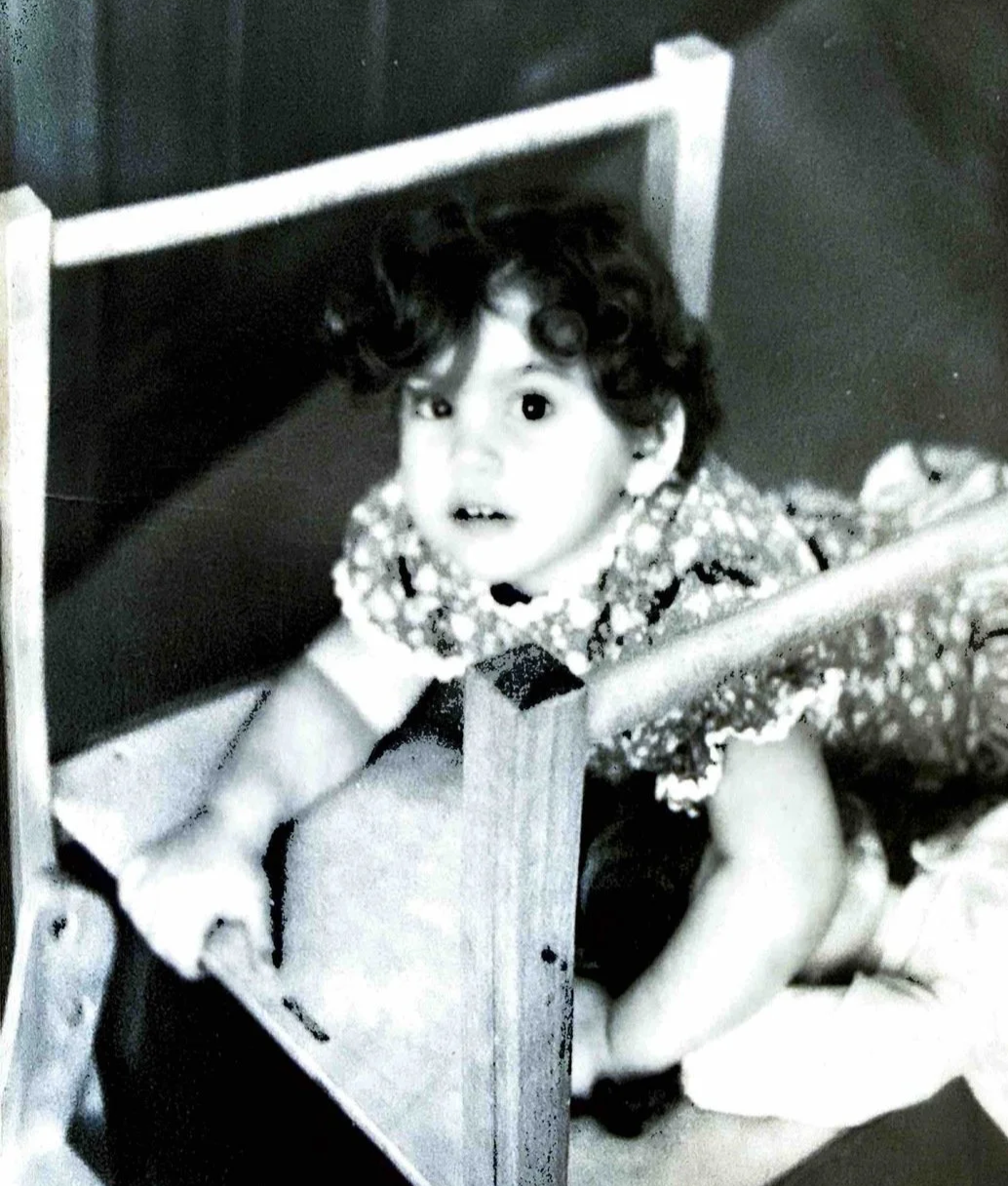 A young child with curly hair peeking up from behind a wooden piece of furniture, possibly a small table or bed frame, while lying on the floor.