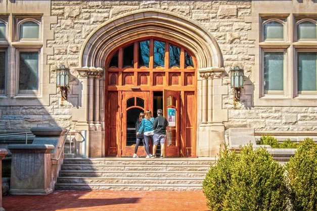 Three people standing at the entrance of a large stone building with a wooden door and arched window, surrounded by steps and bushes.