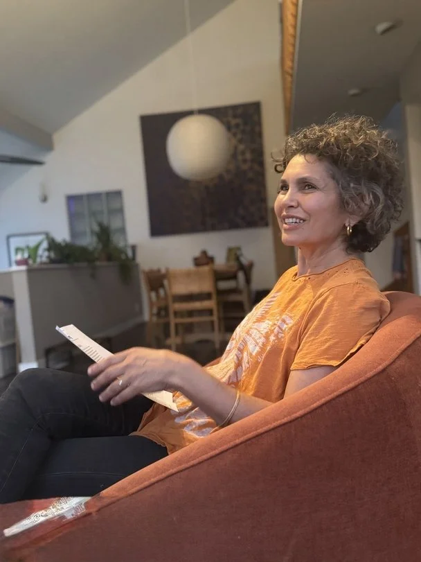 A woman with curly gray hair, wearing an orange T-shirt, sitting on a reddish brown couch in a living room, smiling and holding a piece of paper or a receipt in her hand.