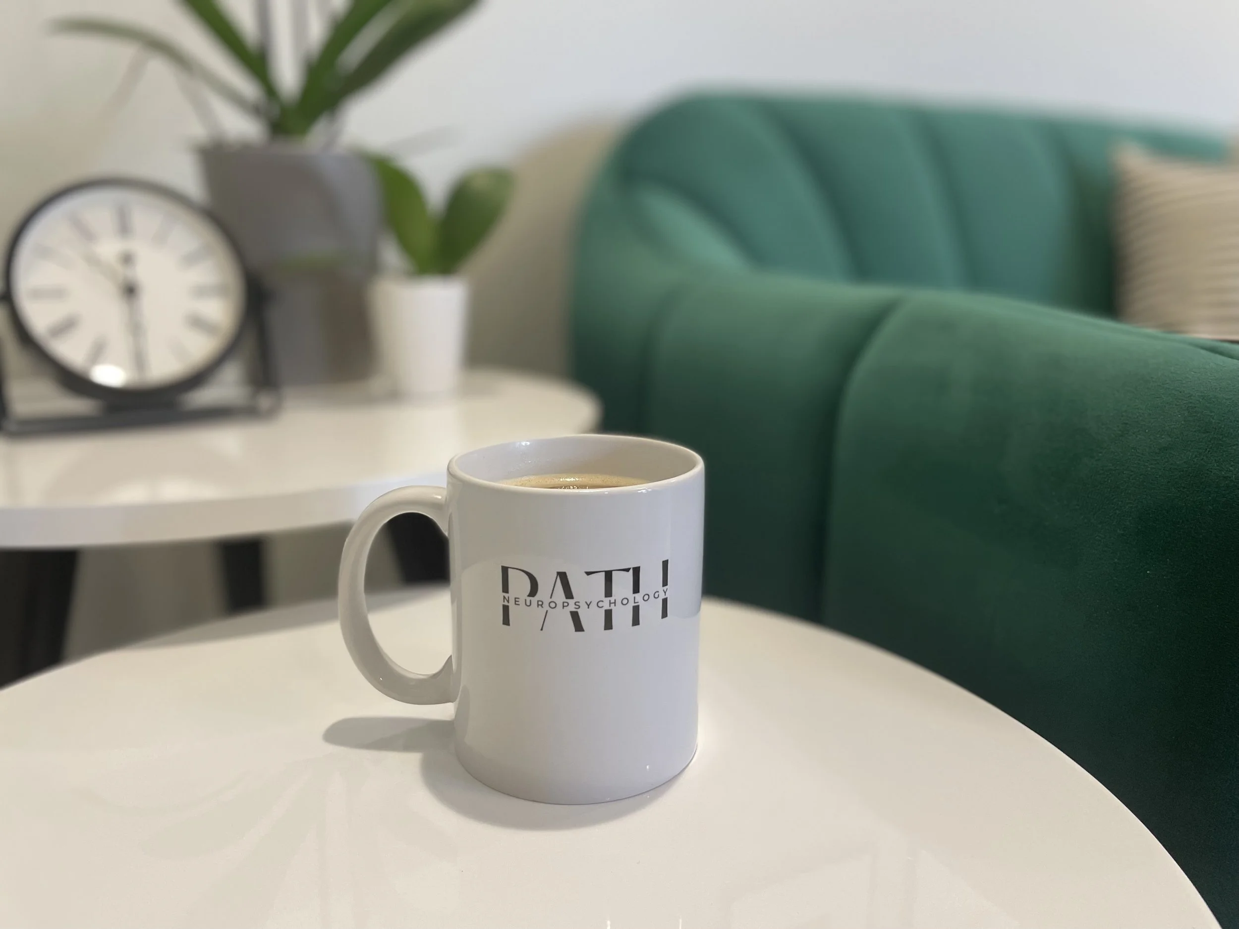 White coffee mug with black text on a white table, with a green couch in the background and a white side table with a clock and potted plant.