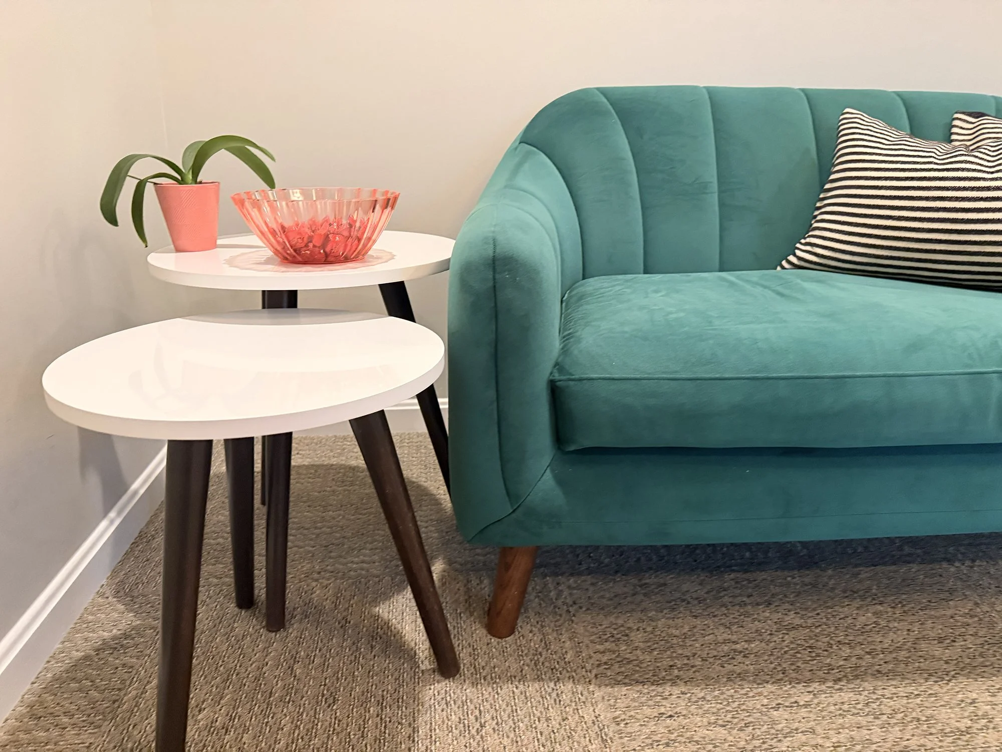 PATH Neuropsychology office with a teal upholstered sofa, a black and white striped cushion, two white tiered side tables, a pink pot with a green plant, and a pink glass bowl on the tables.