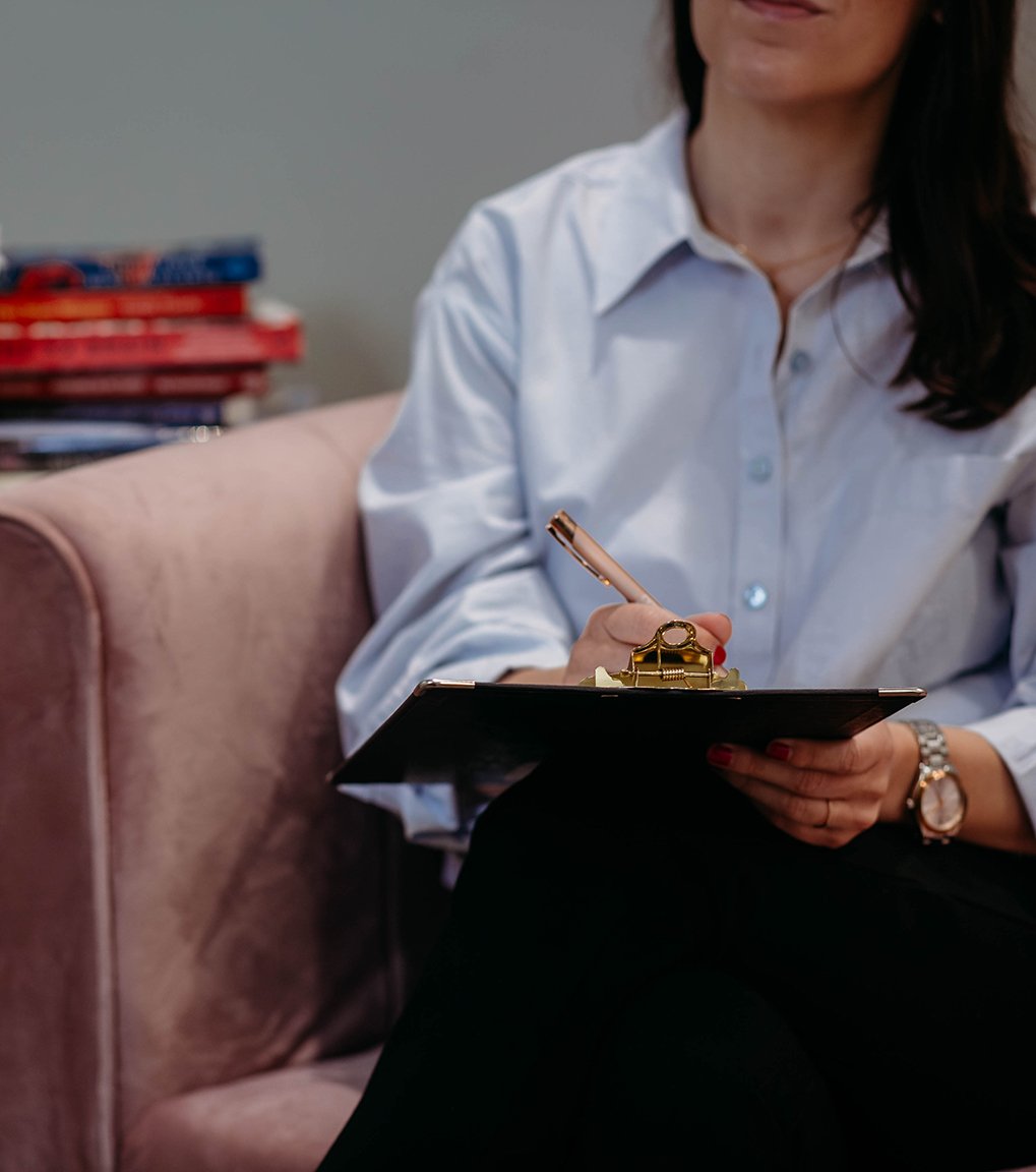 A woman in a white shirt sitting on a pink sofa, holding a black clipboard and writing with a gold pen, with a watch on her wrist and a pile of red and blue books in the background.
