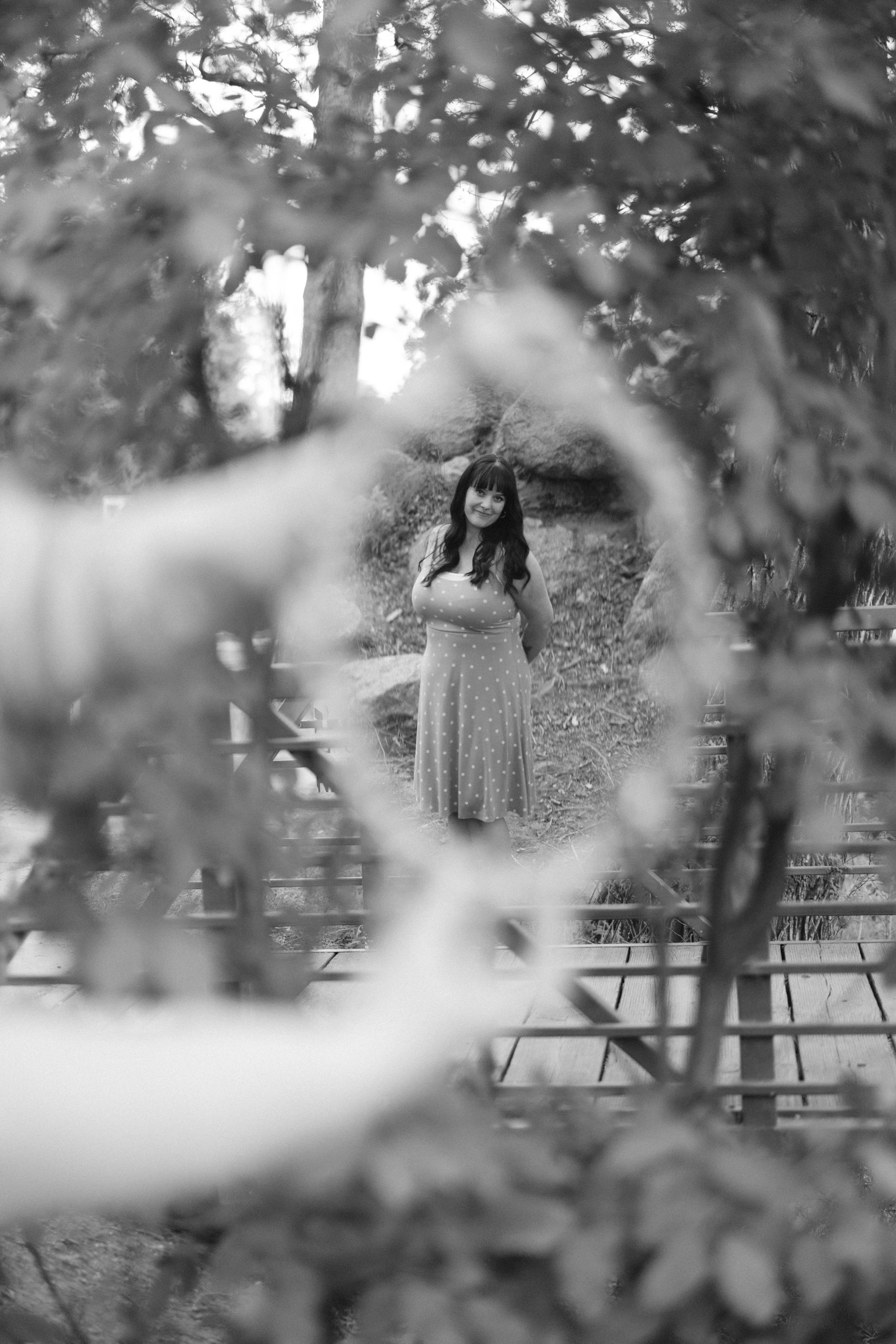 A woman in a polka dot dress standing outdoors, framed by blurry leaves and branches in the foreground, with trees and rocks in the background.