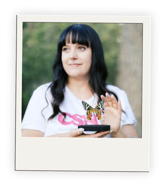A woman with black hair and bangs holding a butterfly on her hand outdoors.