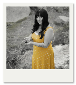 A woman in a yellow polka dot dress standing near a stream, smiling at the camera.